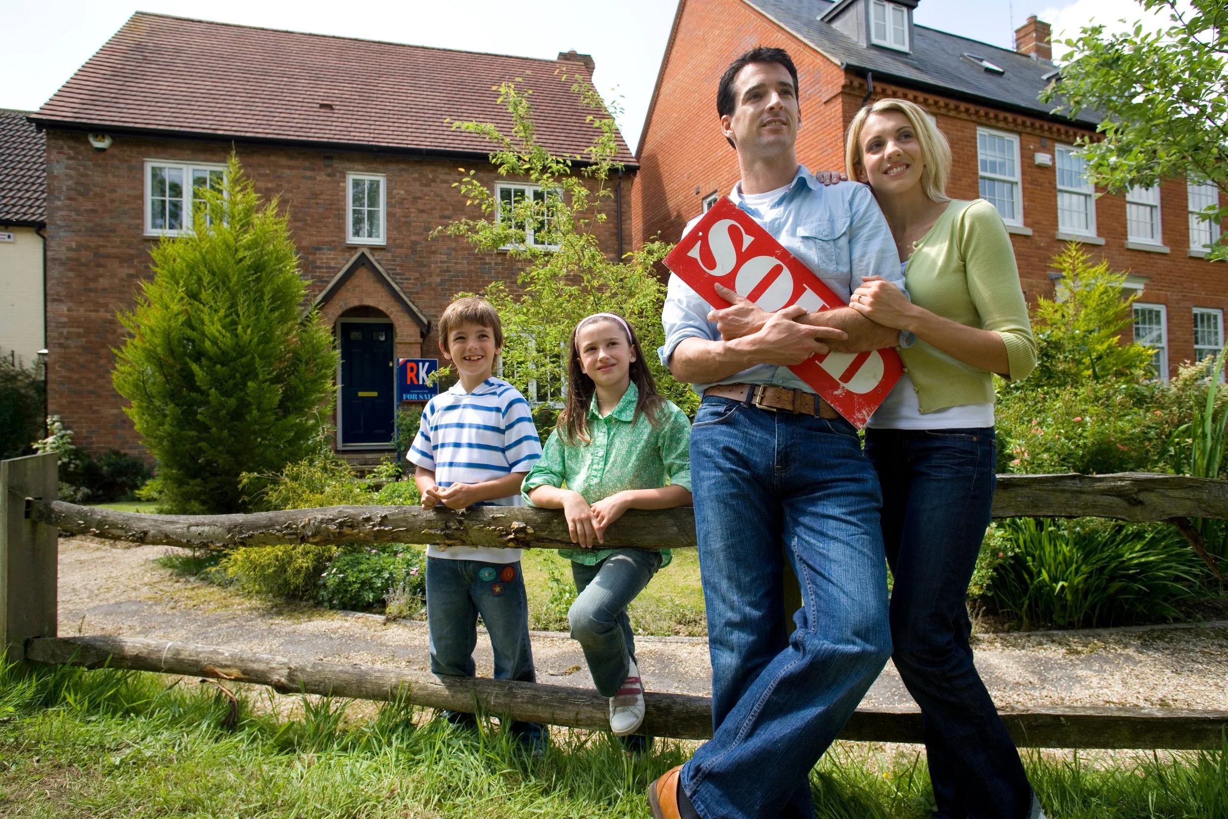 A happy family of four standing in front of a house for sale, with a real estate agent holding a 'Sold' sign. The family includes a man, a woman, and two children, a boy and a girl, all smiling and looking at the camera, with greenery and a wooden fence in the background.
