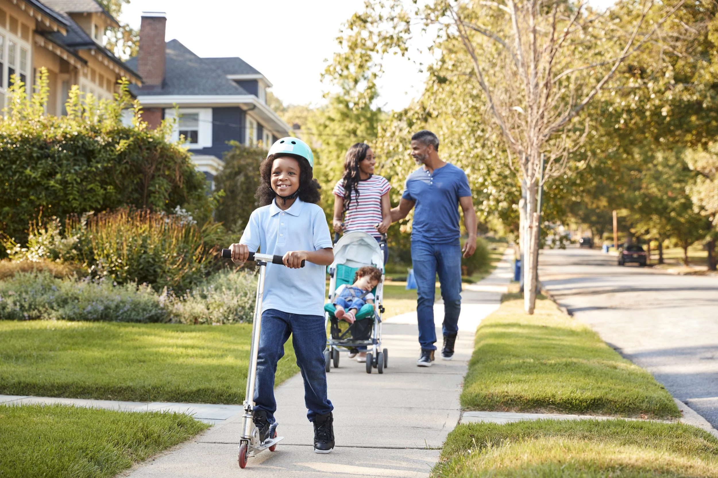 A young girl wearing a helmet rides a scooter on a suburban sidewalk while her family walks behind her. A woman and a man walk with a girl and a sleeping baby in a stroller. The scene is set in a tree-lined neighborhood during daytime in autumn.