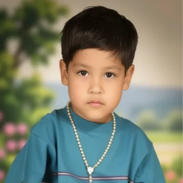 Young boy with short black hair wearing a blue t-shirt and a pearl necklace, standing outdoors with a blurred greenery background.