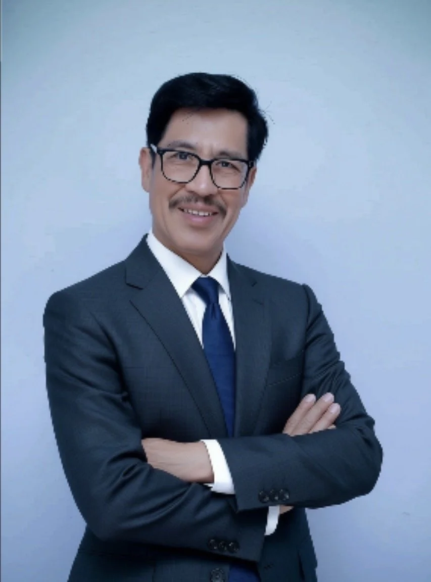 Man in a business suit and glasses with arms crossed, smiling against a plain background.