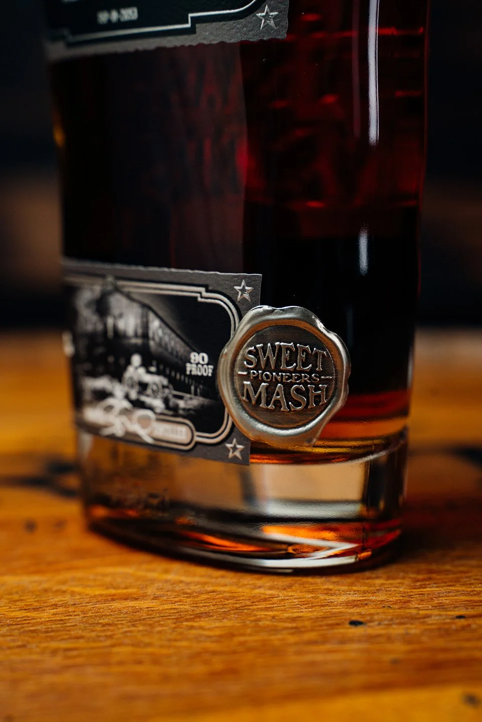 A glass of dark amber liquor with a metal emblem reading 'Sweet Pioneers Mash' and a black and white tag attached, sitting on a wooden surface.