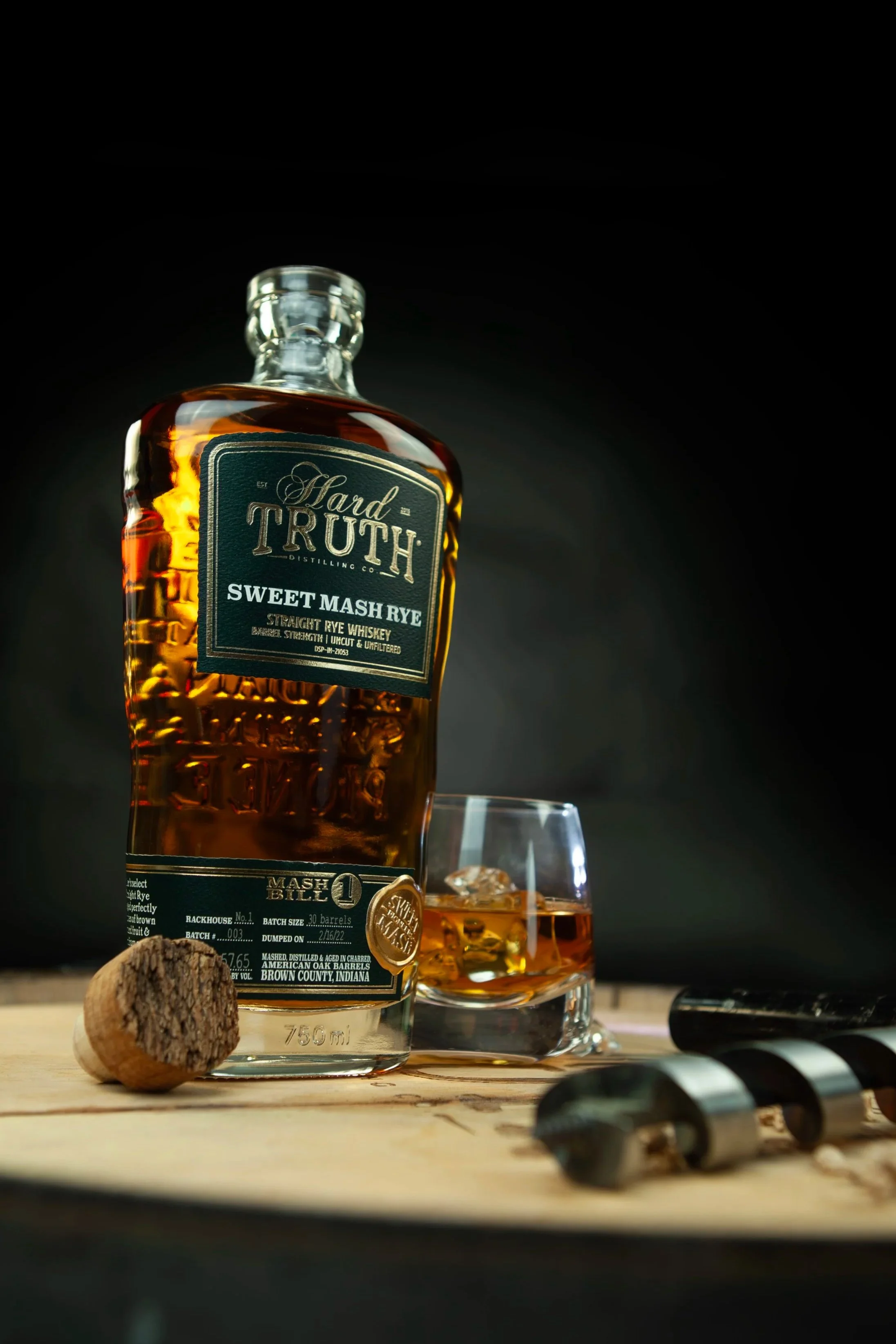 A bottle of Hard Truth Sweet Mash Rye whiskey, a glass of whiskey with ice, a cork, and a corkscrew on a wooden surface against a dark background.