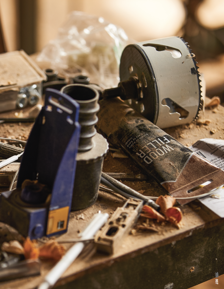 Close-up of a cluttered workbench with power tools, a can of wood filler, electrical wires, and various small items scattered around.