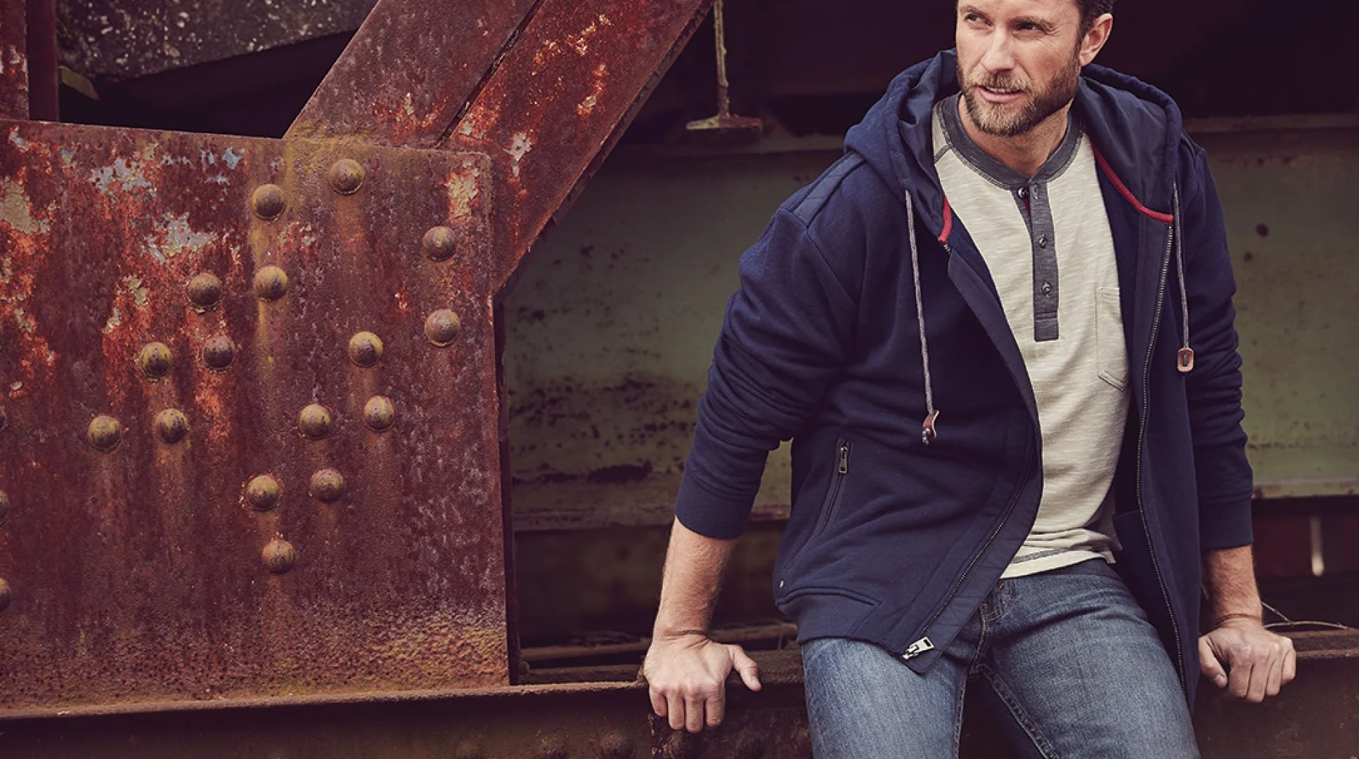 A man in casual clothes sitting on a rusted metal structure outdoors.