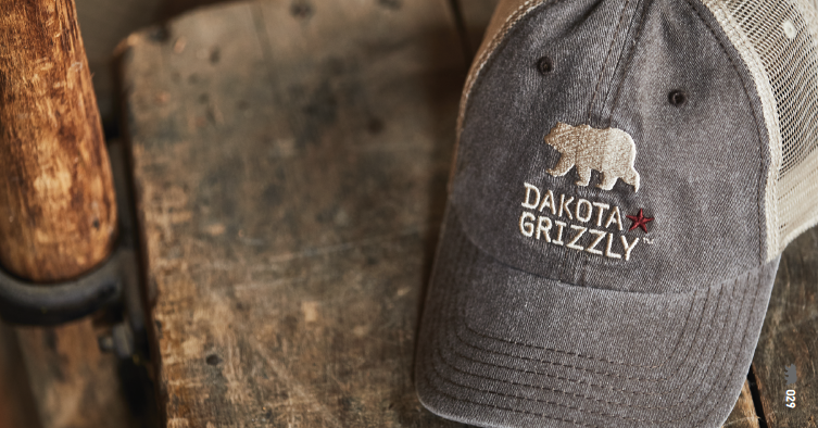 Gray baseball cap with embroidered bear and the words 'Dakota Gritzy' on a wooden surface.