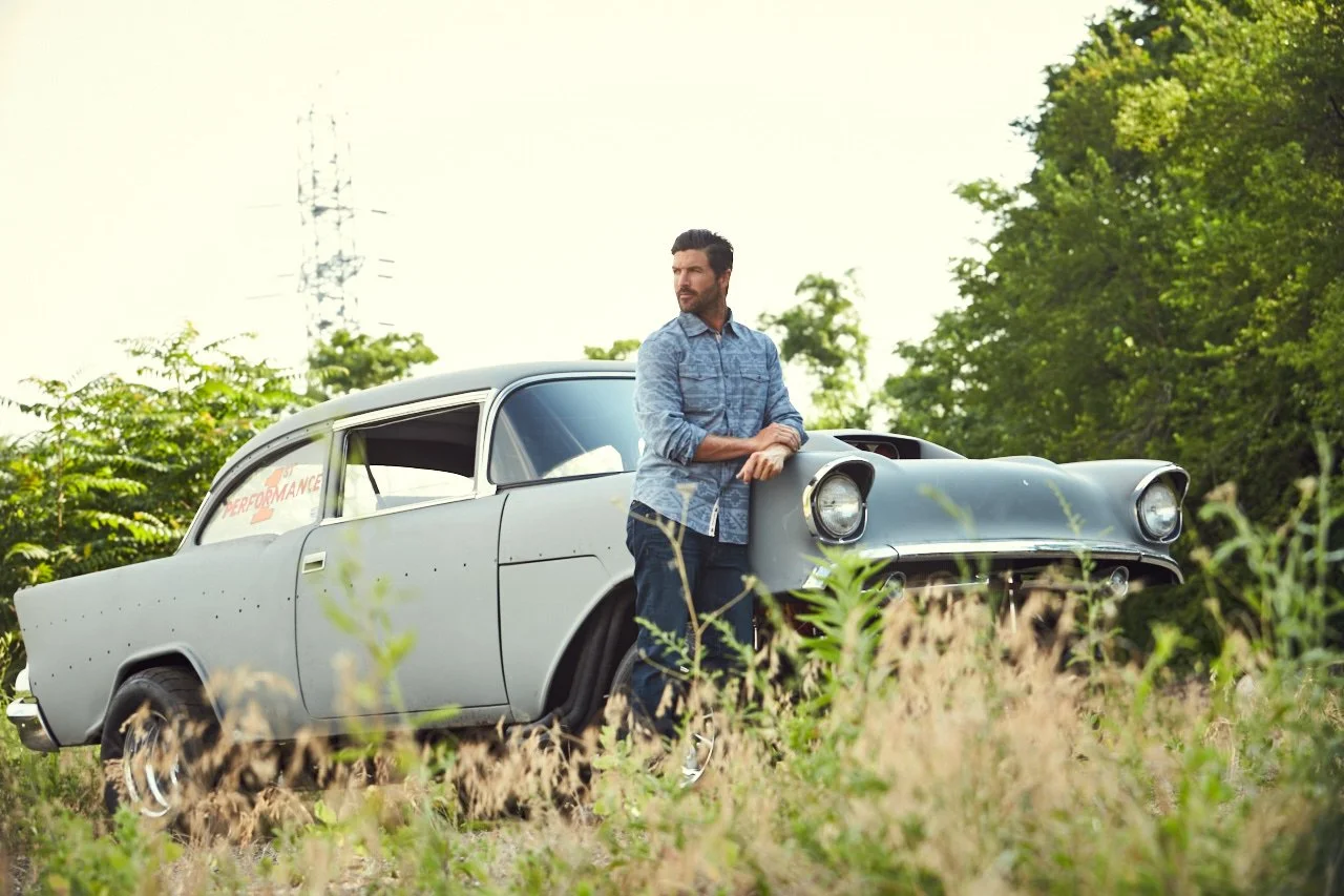 A man leaning on a vintage gray car with a grassy field and green trees in the background.