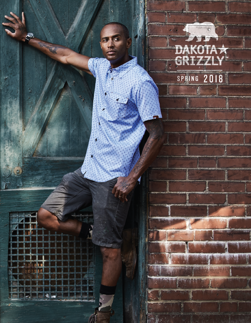 A man standing against a green wooden door with his arm outstretched, wearing a light blue patterned short-sleeve shirt, shorts, and boots, with tattoos visible on his arm. There is a brick wall and a sign that reads "Dakota Grizzly Spring 2018."