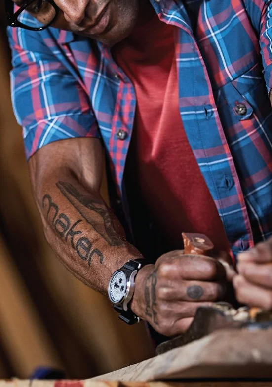 A man with "Maker" tattoos on his arm, wearing a red shirt and blue plaid button-up shirt, is working with tools at a workbench, wearing a wristwatch on his left wrist.