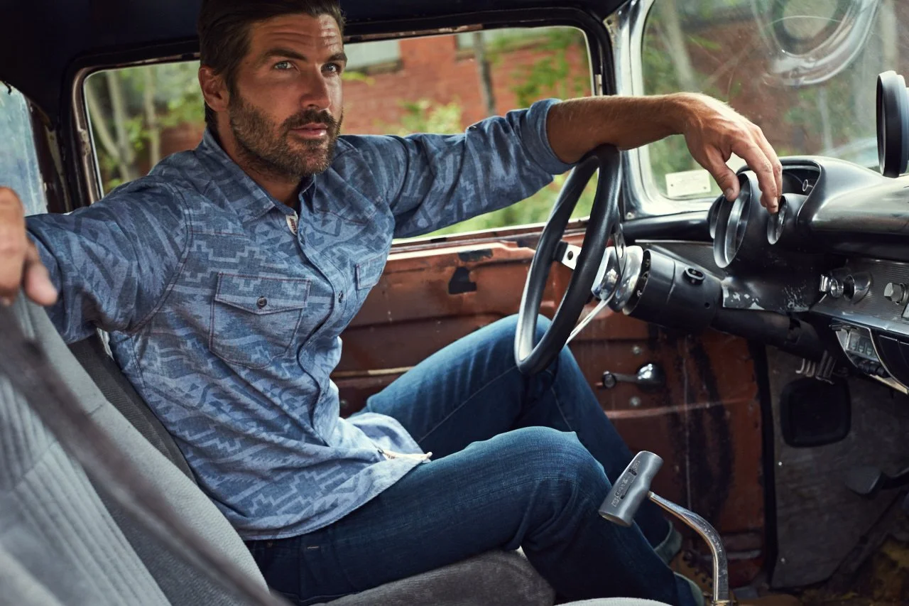 A man with a beard and wearing a patterned blue shirt sitting in the driver's seat of a vintage truck, holding the steering wheel with one hand and looking towards the camera. The interior of the truck is rustic with visible wear.