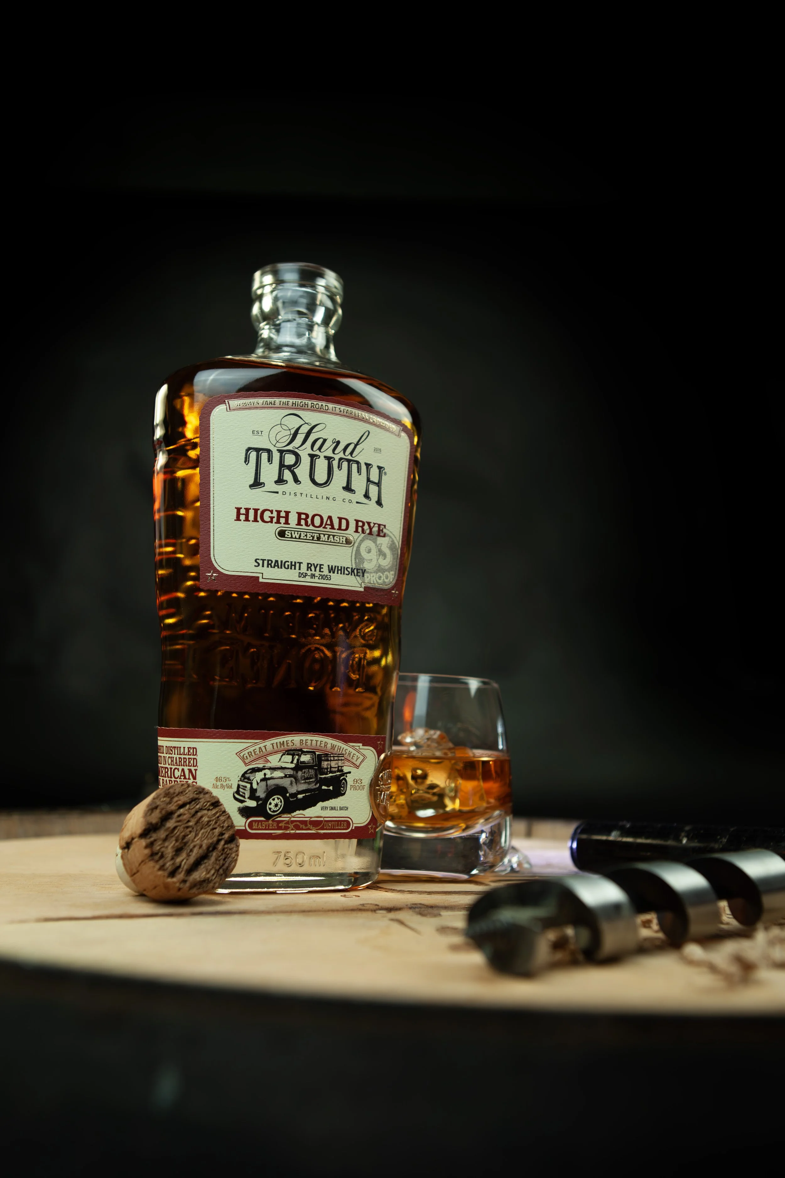 A bottle of Hard Truth High Road Rye whiskey next to a glass filled with whiskey and ice, placed on a wooden surface with a cork and a metallic screw cap nearby, against a dark background.