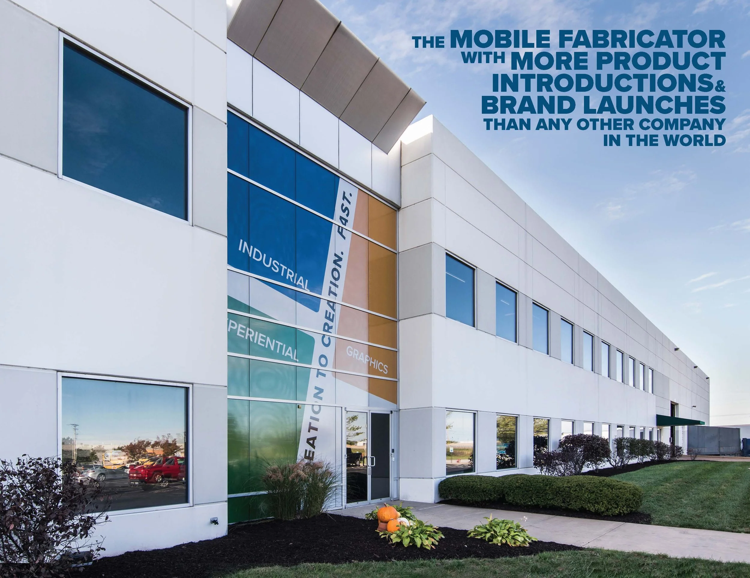 Exterior of a modern white commercial building with large tinted windows, landscaped bushes, pumpkins, and plants in front. A large sign with colorful segments on the wall says 'Industrial, Experimental, Creative, Graphics, Fast.' Blue sky with some 