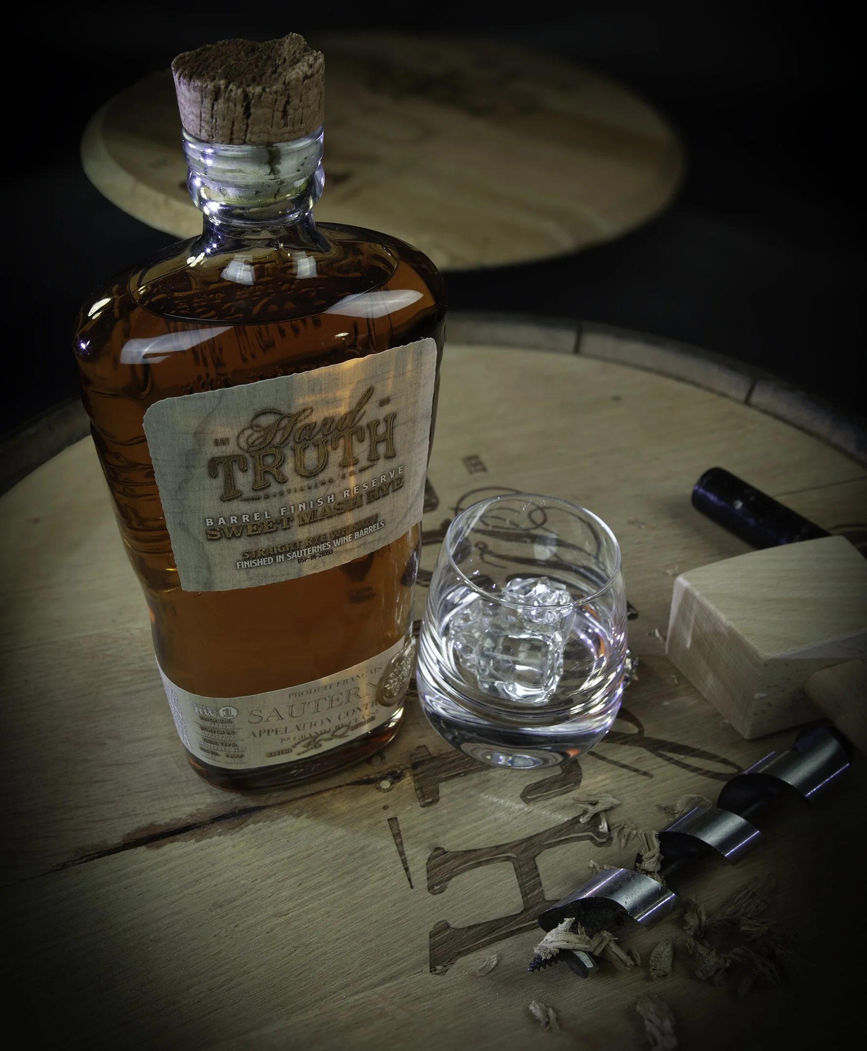A bottle of whiskey labeled 'Hard Truth' next to a glass with ice on a wooden table with wine barrel markings, surrounded by a small wooden block, a black object, and a corkscrew.