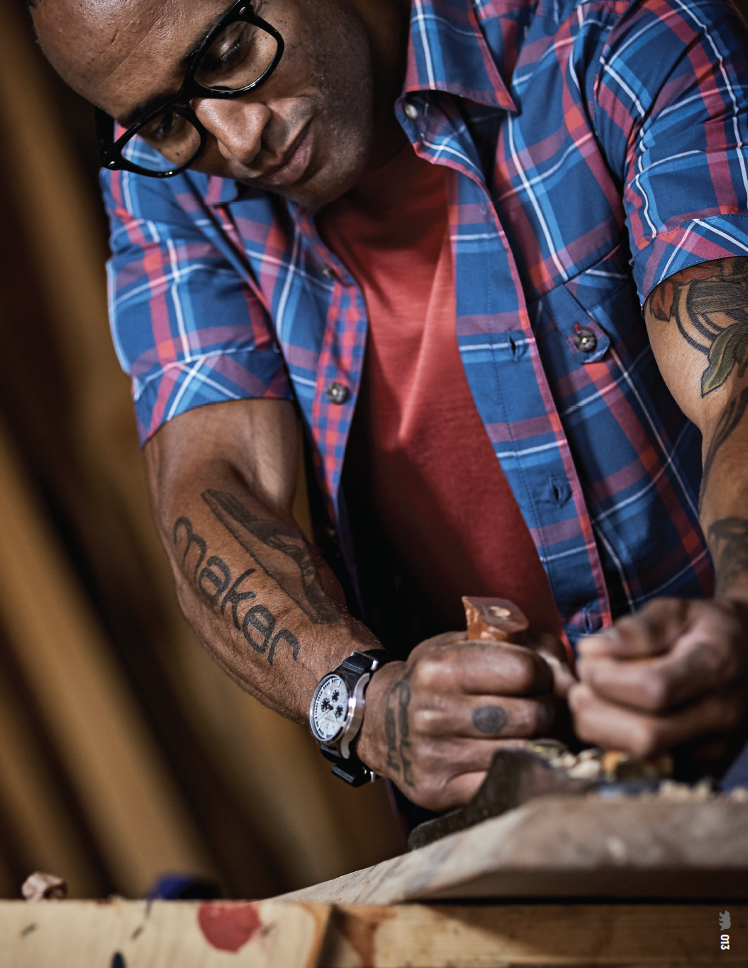 A man with glasses and tattoos on his arms carving or crafting something on a wooden surface in a workshop.