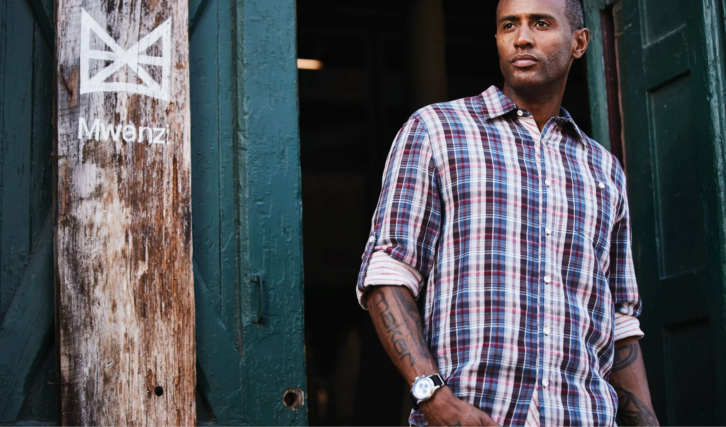 A man with tattoos on his arms wearing a plaid shirt standing at a rustic wooden and green painted building.