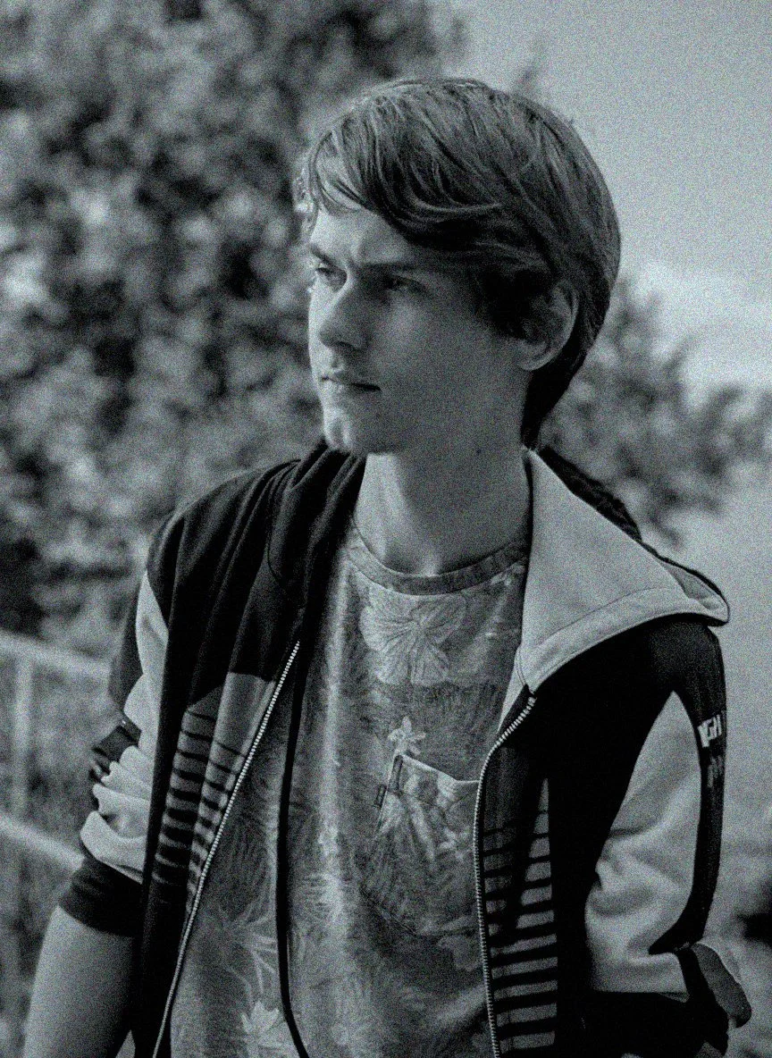 Black and white photo of a teenage boy with short, layered hair looking to the side outdoors, wearing a patterned shirt and a zip-up jacket with stripes.