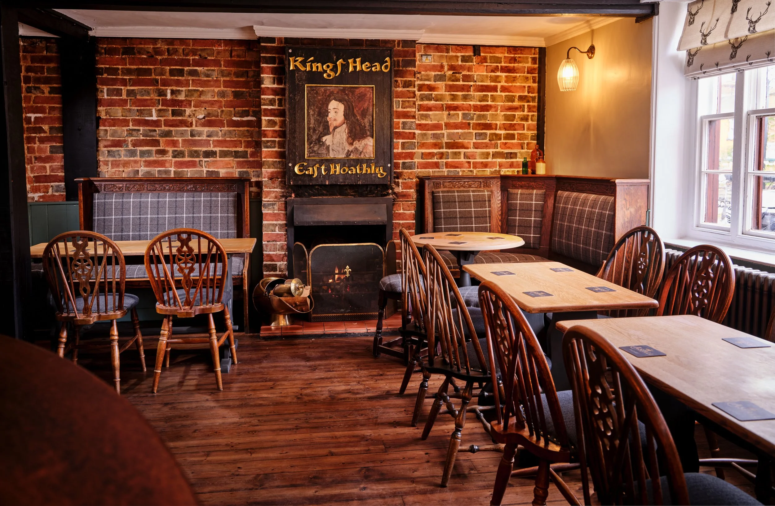 Interior of a cozy pub with wooden tables and chairs, exposed brick wall, and a fireplace, called "King's Head" with a portrait of a king and the text "Ear Hoathing."