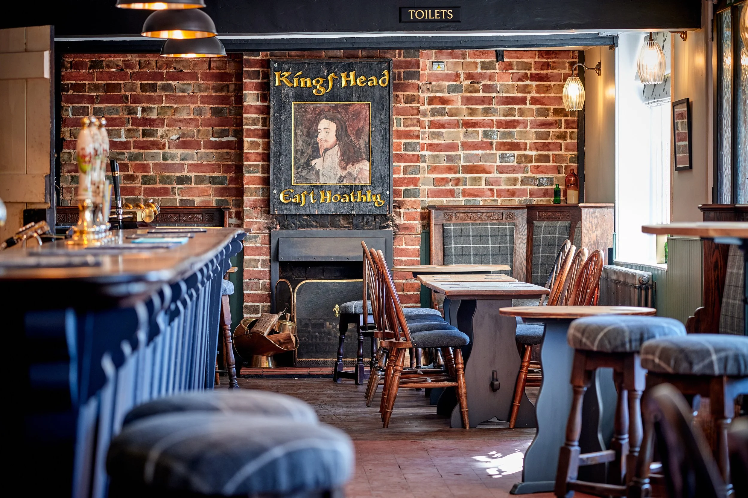 Interior of a cozy pub or restaurant with brick walls, wooden tables and chairs, paintings, and warm lighting. A sign indicates the restrooms nearby.