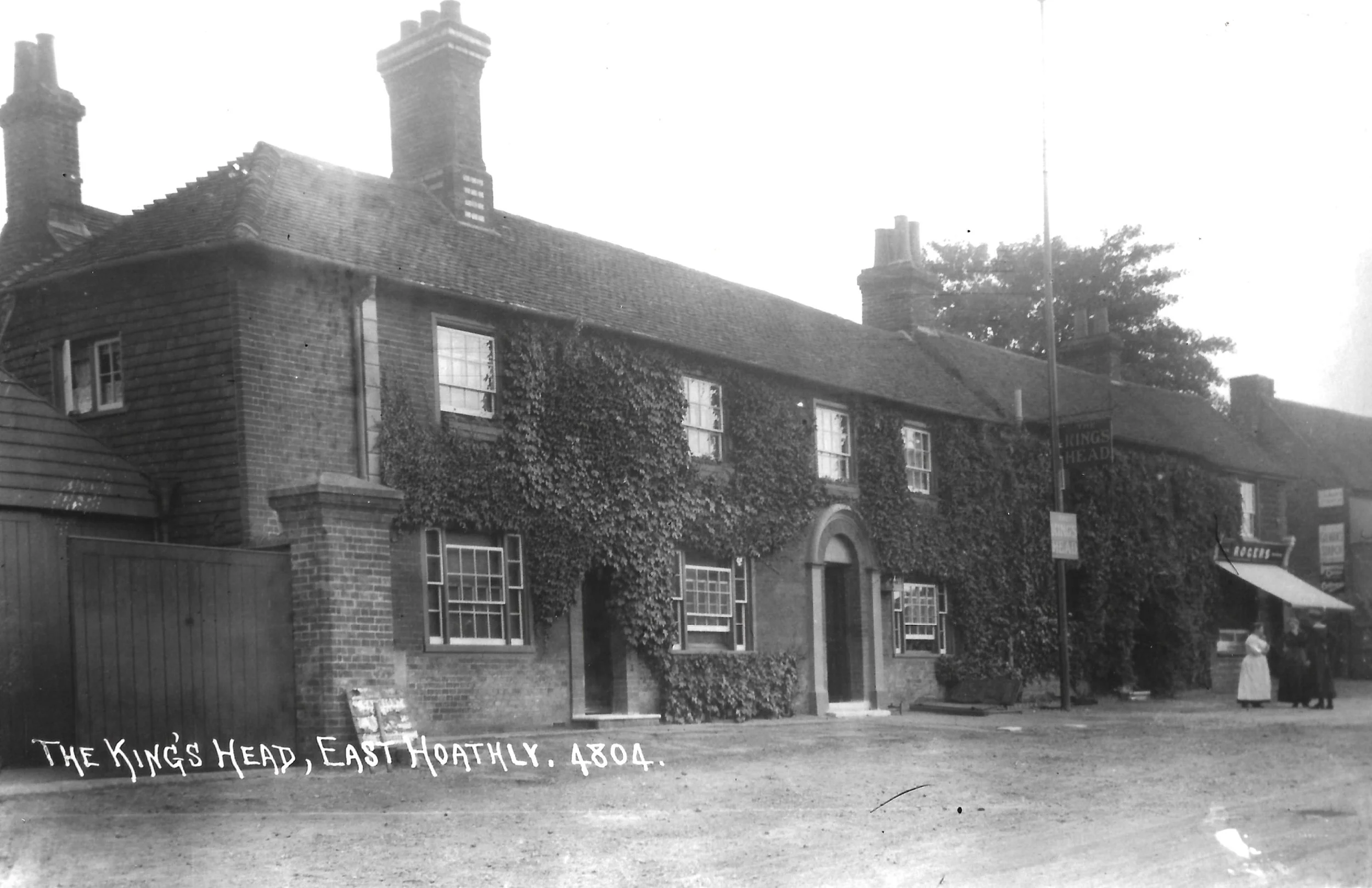 Black and white photograph of a brick building covered in ivy with multiple chimneys, labeled as The King's Head, East Hoathly, 4804, with a sidewalk, street, and people standing near a shop on the right side.