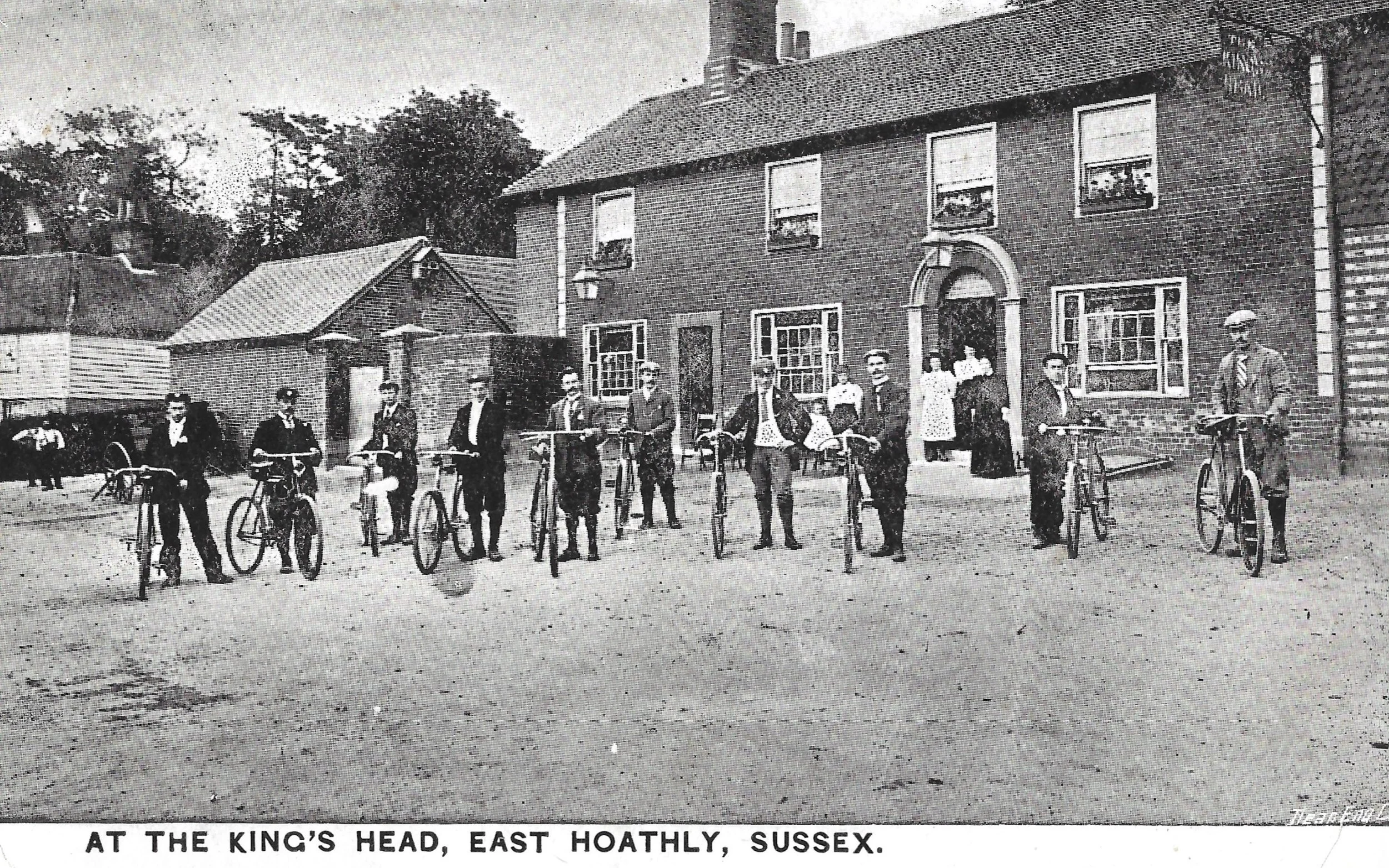 Historical black-and-white photo showing a group of people with bicycles standing in front of a brick building labeled 'The King's Head' in East Hoathly, Sussex, with some individuals standing on the porch and others on the ground, and children in the background.