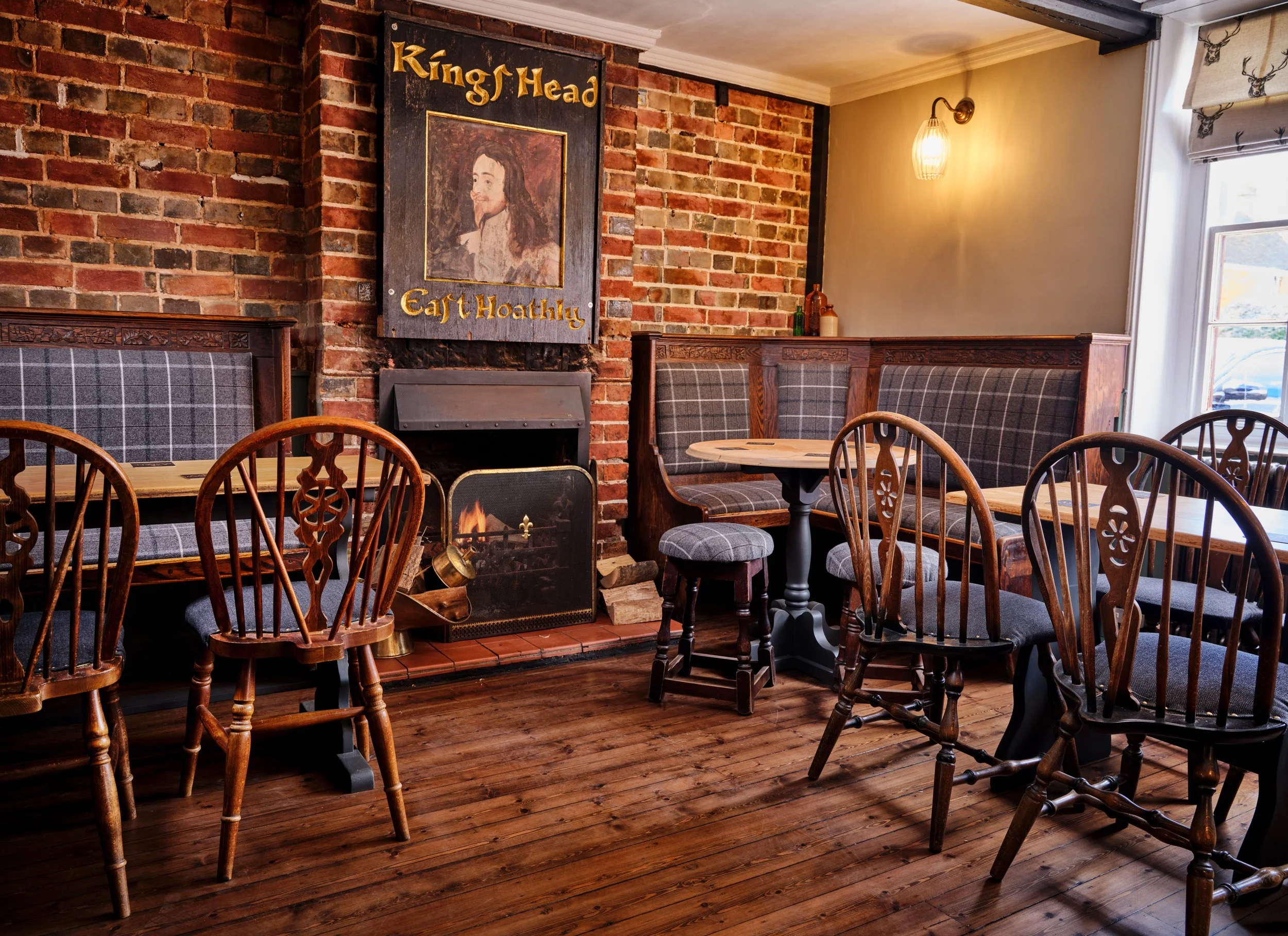 Interior of a cozy pub or restaurant with wooden furniture, brick wall, a fireplace, and decorative sign reading 'King Head' with a portrait of a woman, and a window with a deer head curtain.
