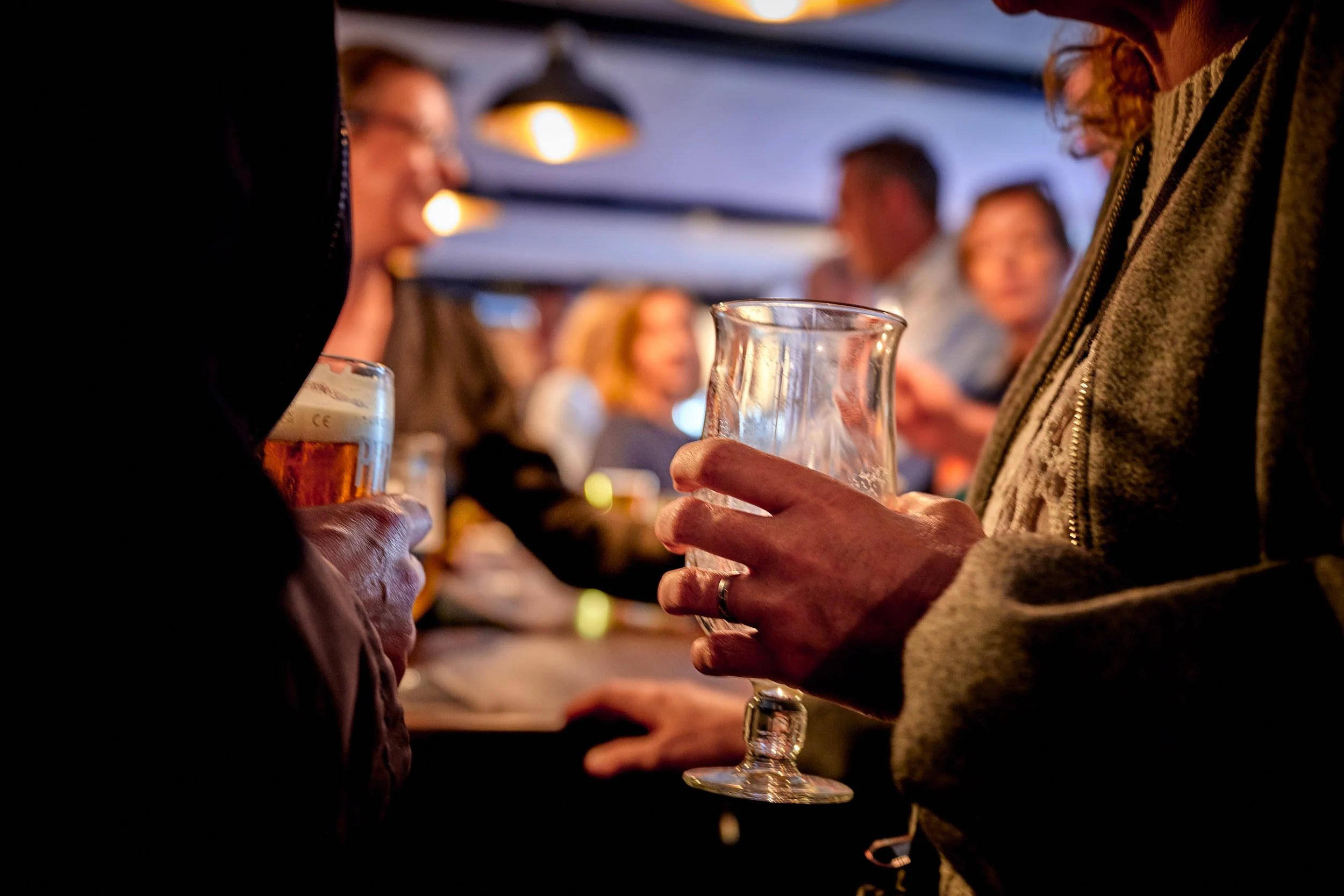 People standing at a bar or social gathering, holding drinks and engaging in conversation. Focus on a person gripping a clear glass with a drink in it, with other individuals blurred in the background.