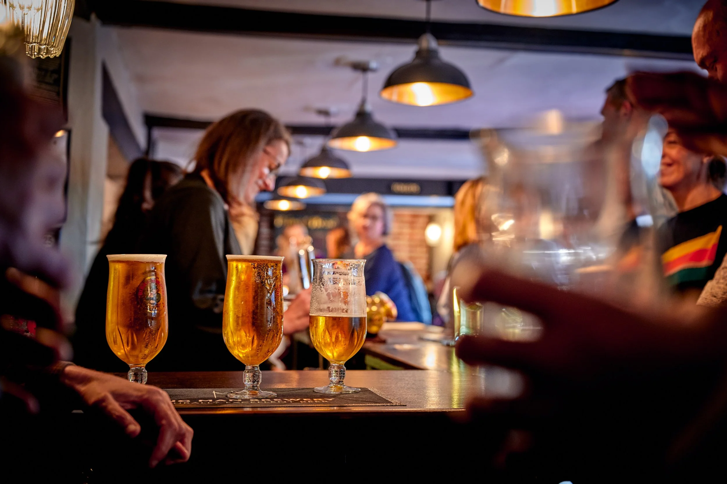 Three glasses of beer on a bar counter in a busy pub, with people socializing in the background.