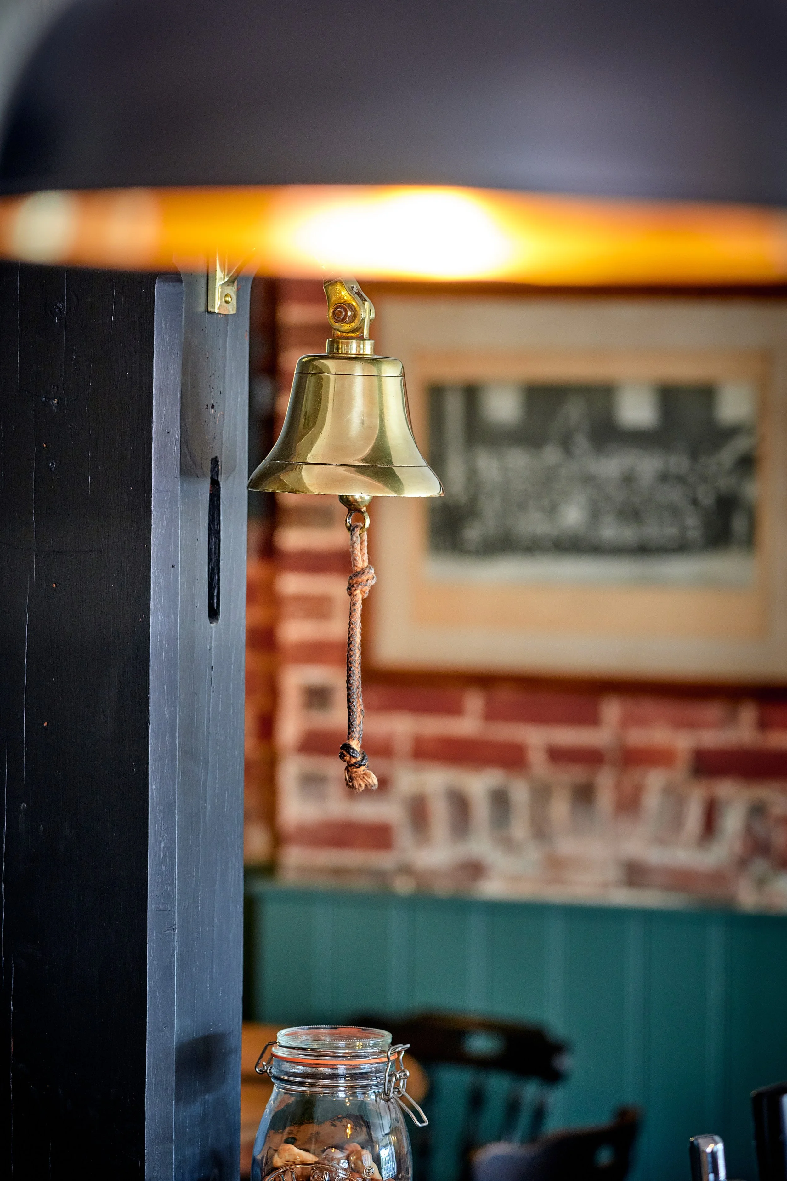 A brass bell hanging from a black wall in a restaurant or cafe, with a blurred background of a brick wall, framed artwork, and a glass jar on a table.