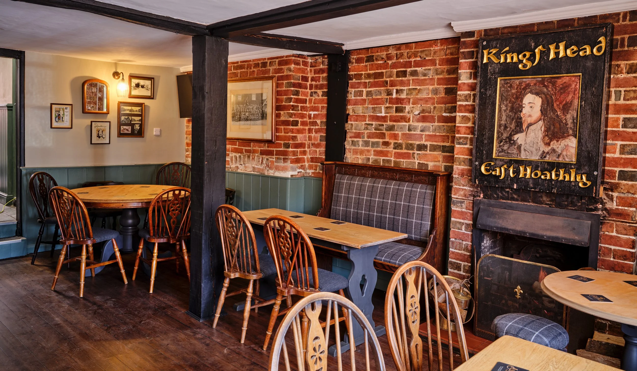Interior of a cozy restaurant with brick walls, wooden tables, and assorted chairs. There is a vintage sign with a portrait and the text 'King's Head' and 'Caff Hoathly' above a fireplace.