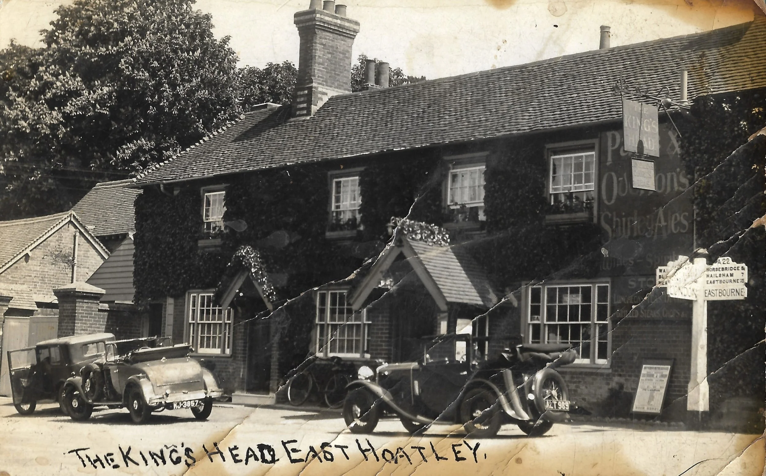 Black and white photo of a two-story brick building with multiple windows and ivy growing on the exterior. Classic cars are parked in front, and there are signs indicating directions to Eastbourne and other places. Text handwritten at the bottom read