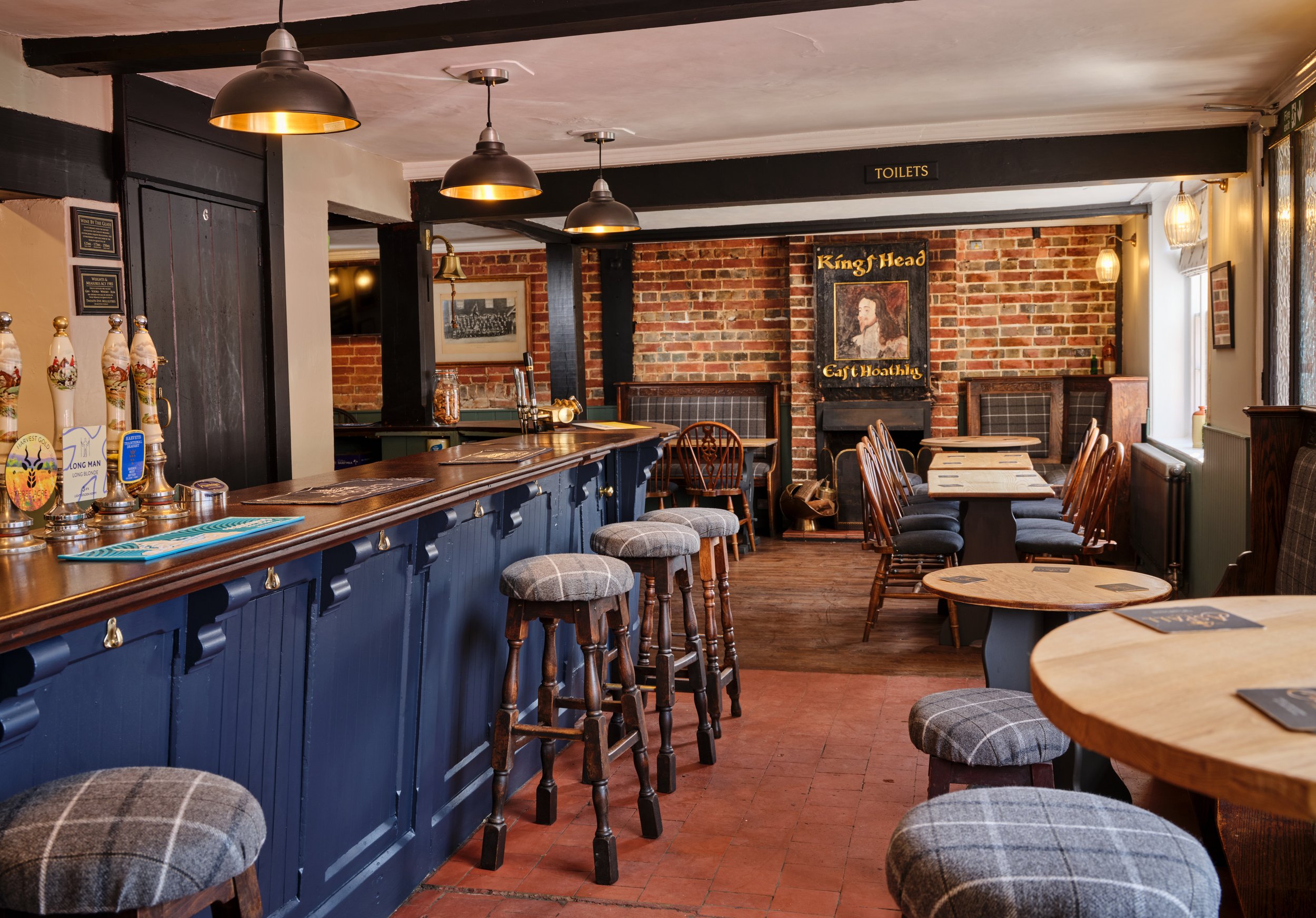 Interior of a cozy pub with a wooden bar, bar stools with plaid cushions, and tables with chairs, brick walls, and vintage decor.