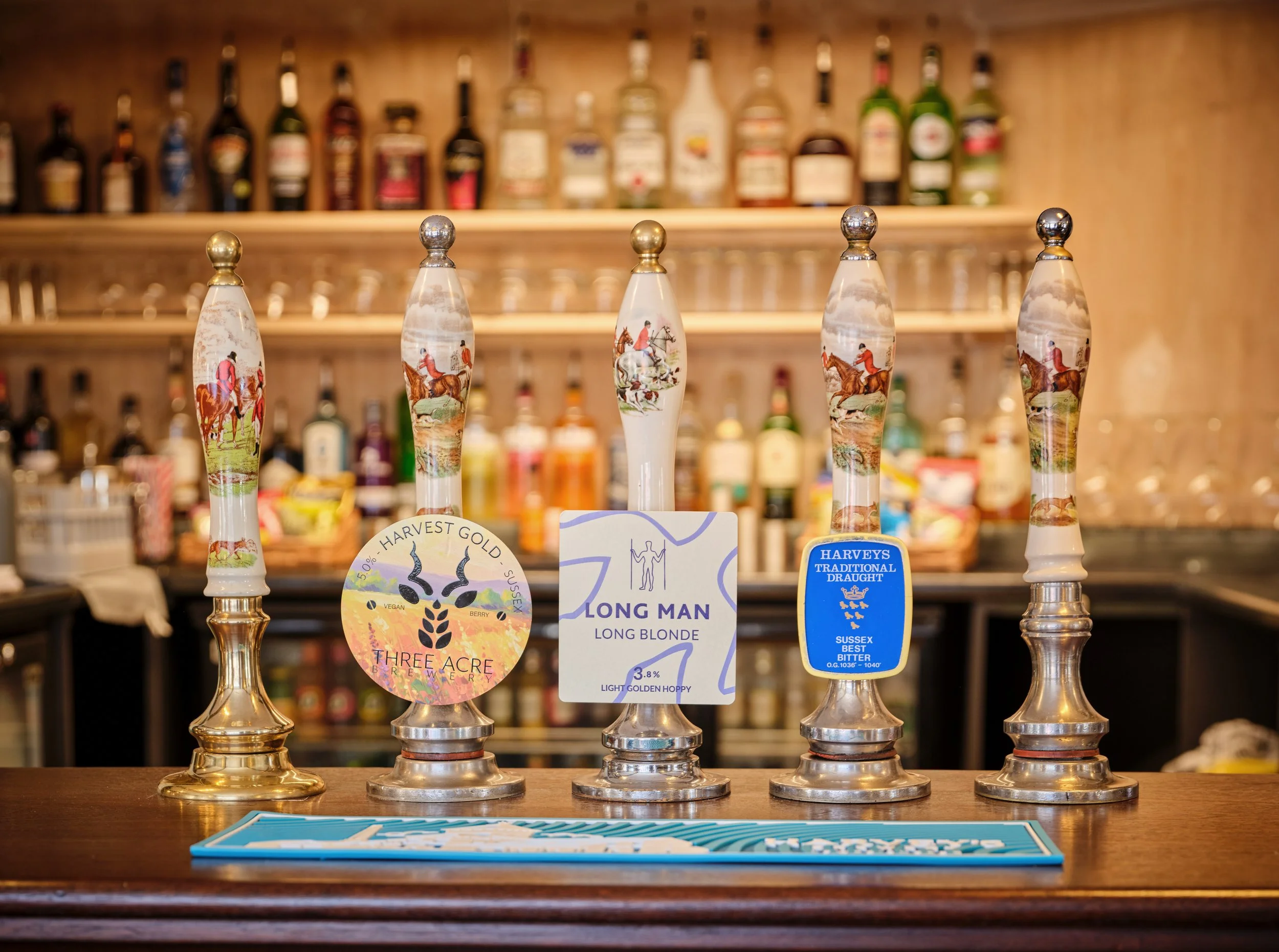 Five beer taps with decorative handles, each featuring a horse rider scene, on a bar counter with behind-the-bar shelves of liquor bottles.