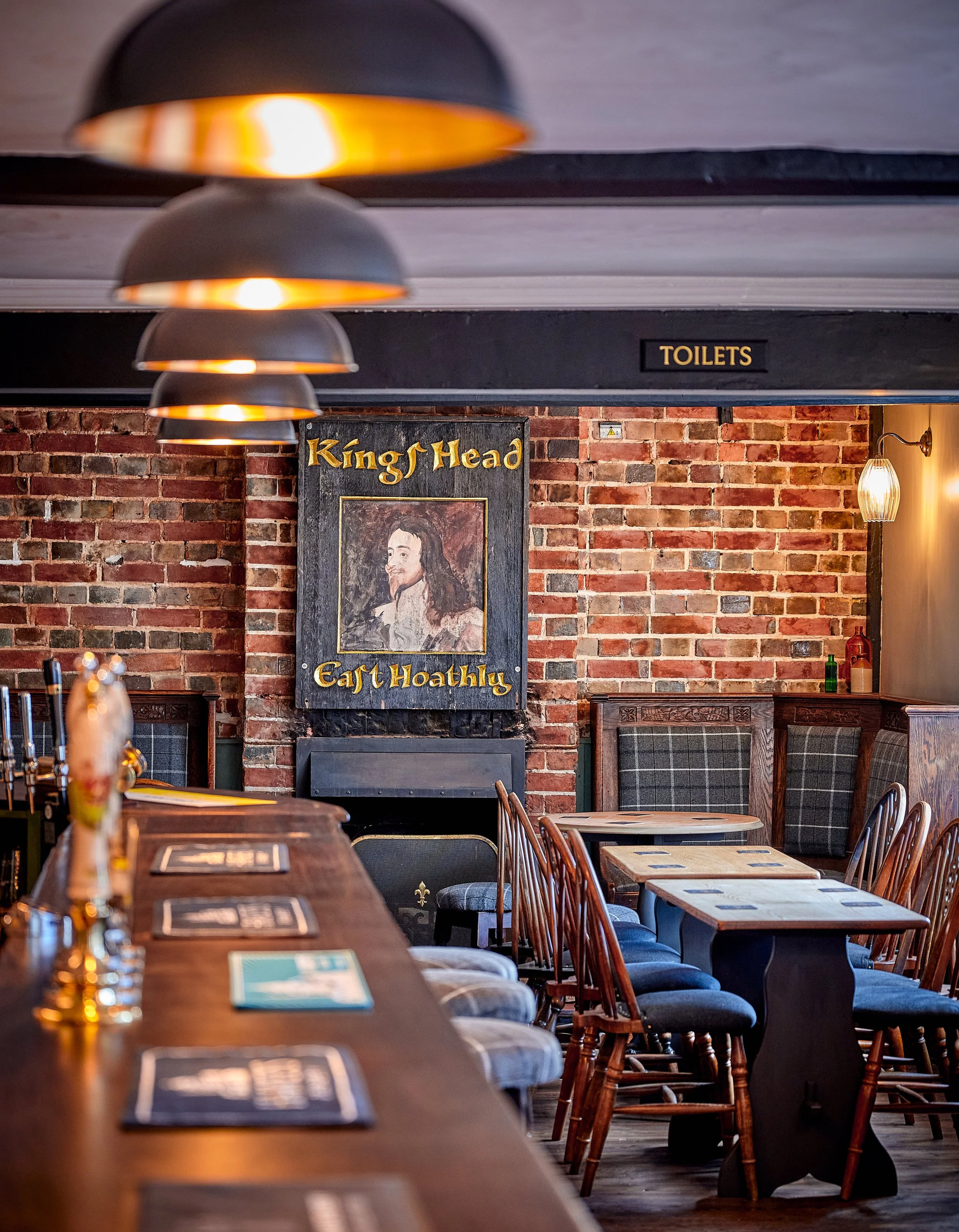 Interior of a pub with wooden tables and chairs, brick walls, and hanging pendant lights. A sign with a portrait and the text "King of Head Eat Hoathy" is visible on the wall. A door labeled "Toilets" is also seen.