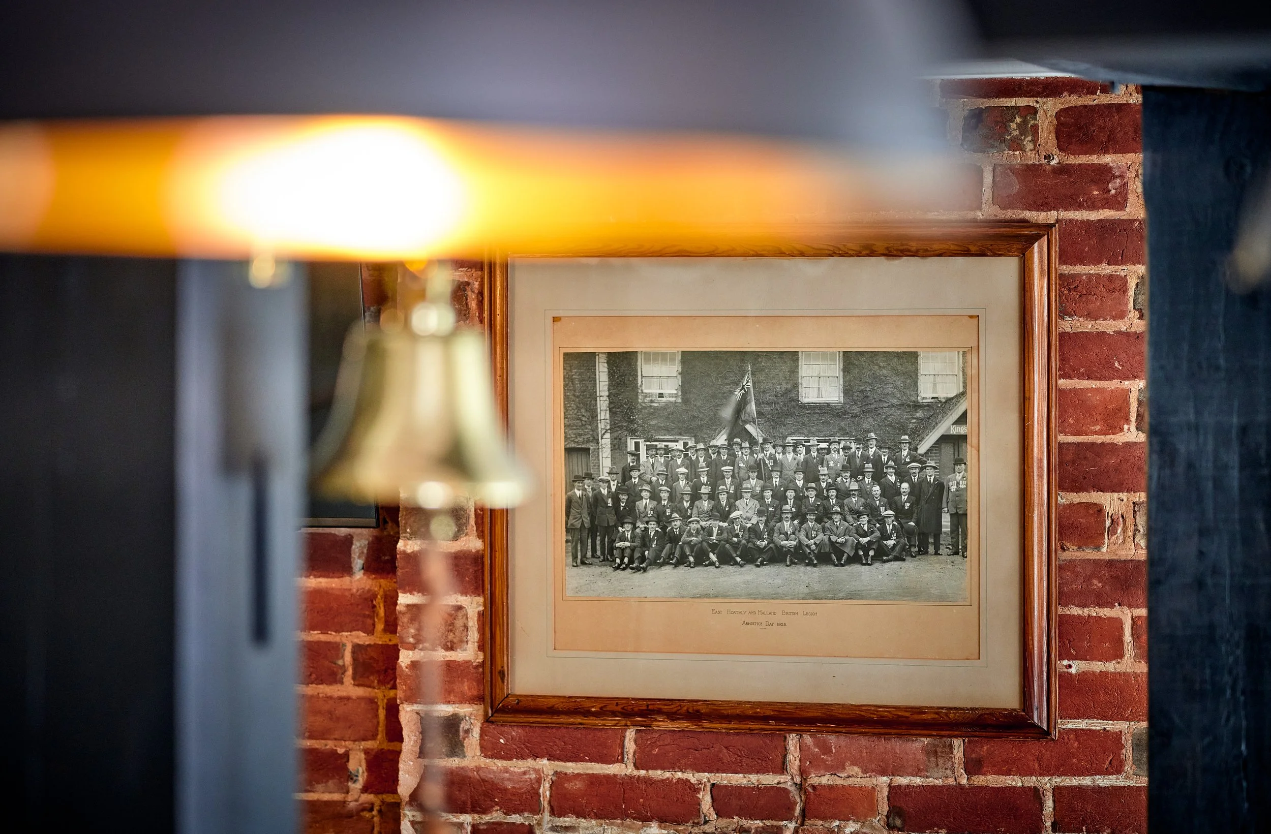 A framed black and white photograph of a large group of men in suits and hats posed outdoors in front of a brick building, with some holding a flag, hanging on a brick wall inside a room.