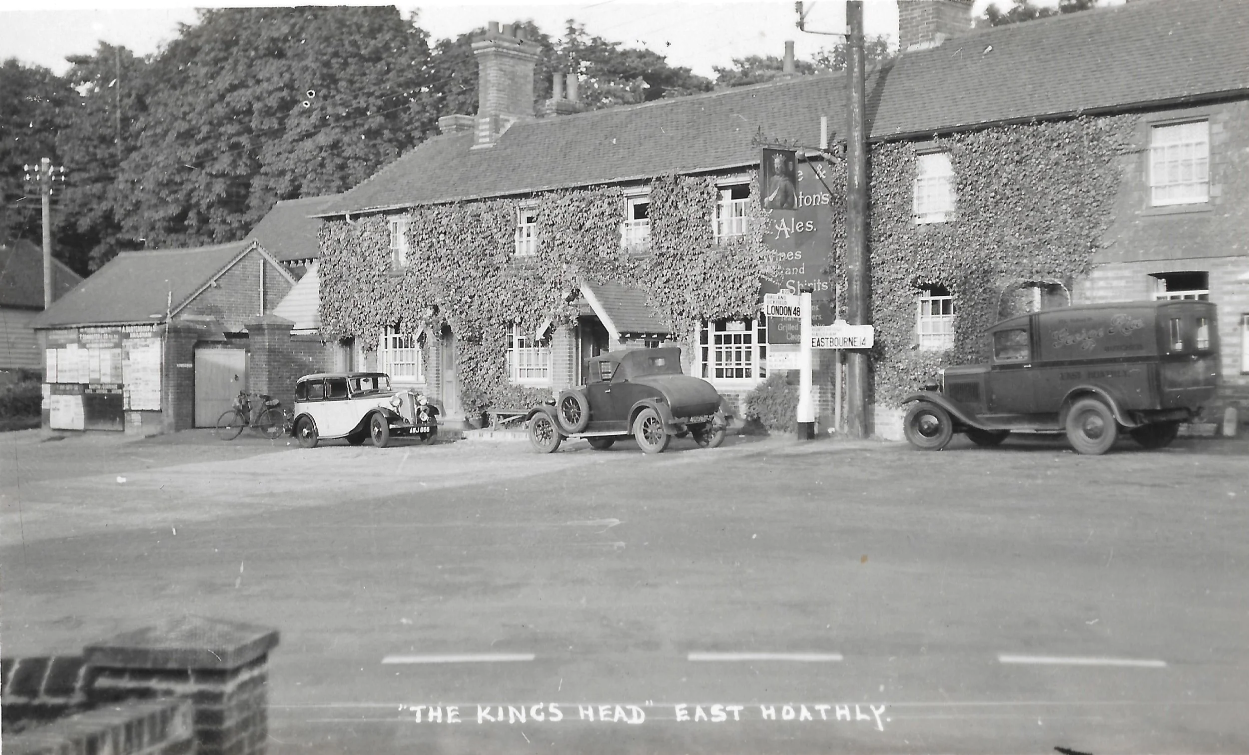 Black and white photo of a traditional pub called 'The Kings Head' in East Hoadly, with ivy-covered walls, three vintage cars parked outside, and a street view with signs indicating directions to London and Eastbourne.