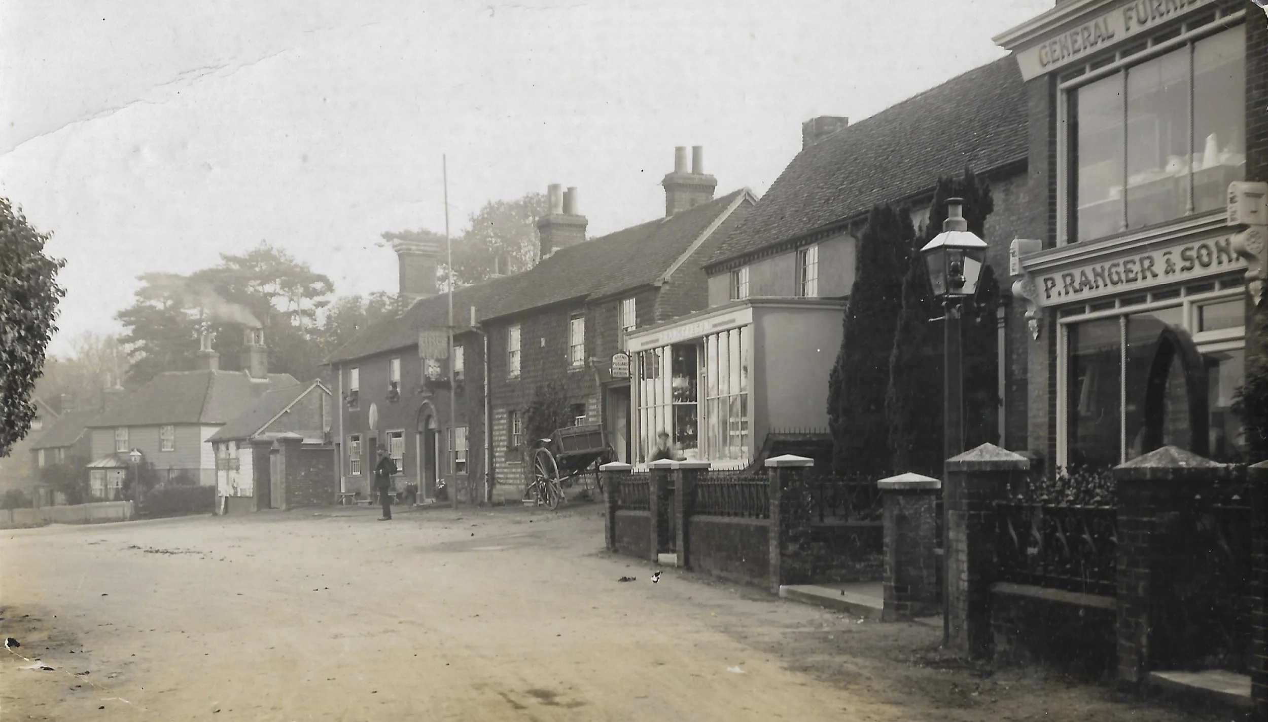 A black and white photograph of a residential street with brick houses, some with chimneys, and a shop with a sign that says 'P. RANGER & SON'. The street is dirt, and there are two men, one standing and one sitting on a cart, visible in the scene. T