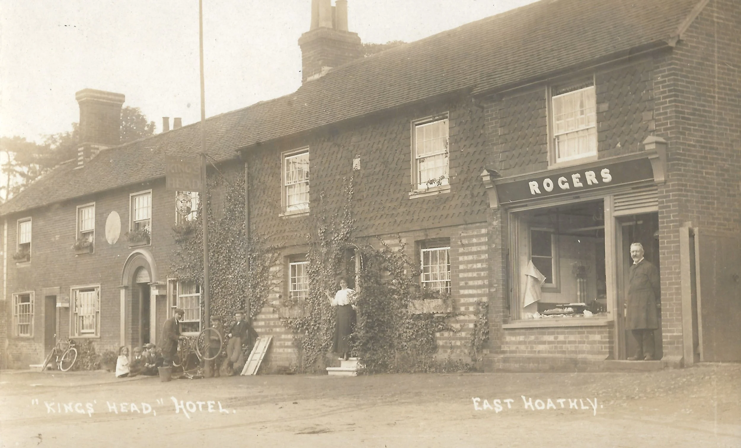 Historical black-and-white photo of a building with a sign that reads 'ROGERS' and people outside. The building is identified as 'King's Head, Hotel' in East Hoathly, with some people standing and sitting on the sidewalk, and a bicycle leaning agains