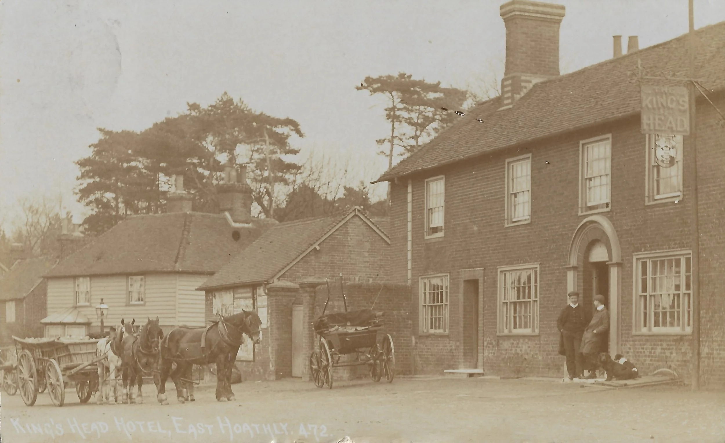 An old sepia-toned photograph of a street scene featuring a brick building labeled 'King's Head' with two men standing outside and a dog lying near the door. There are horse-drawn carts and carriages parked along the street, and trees in the backgrou