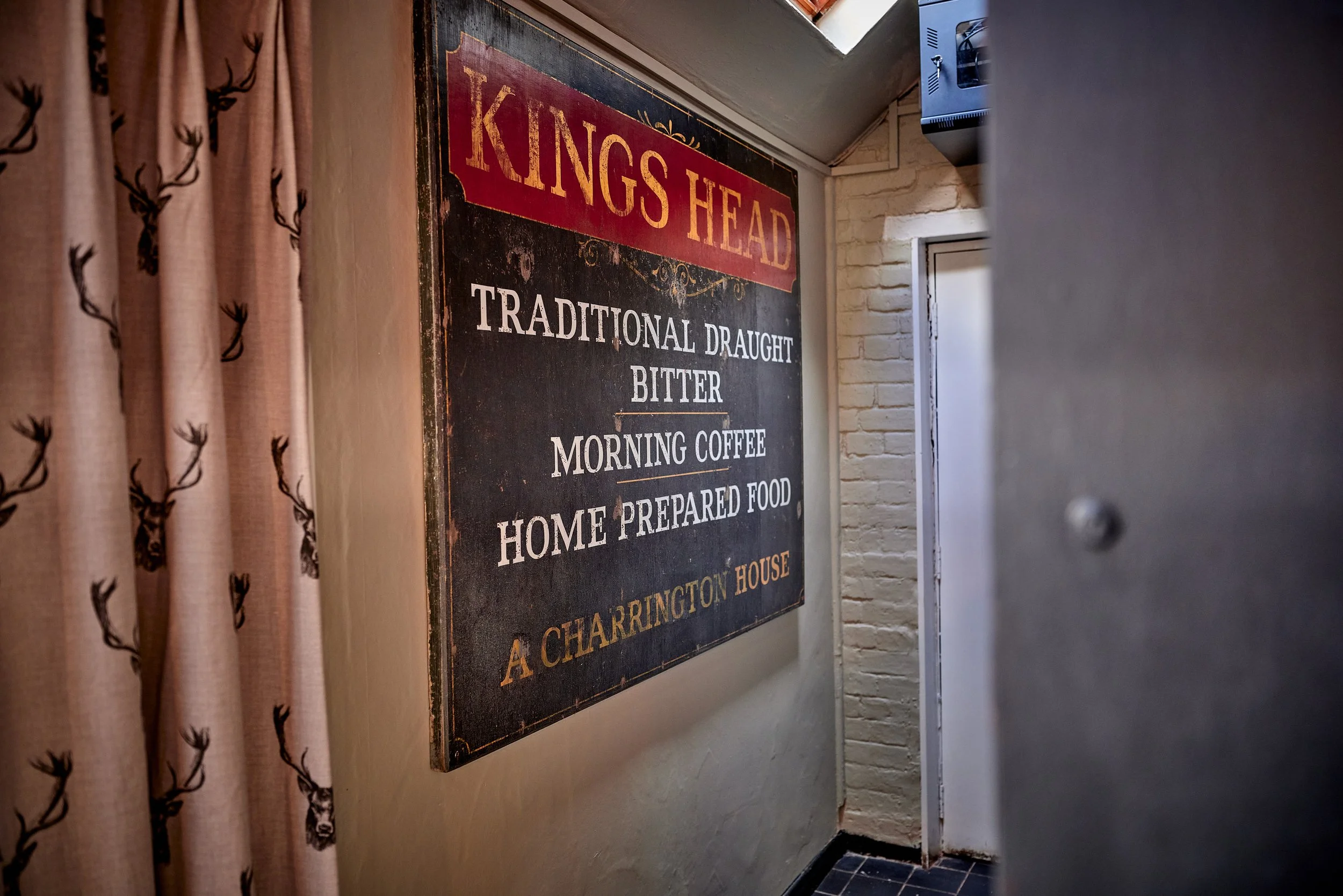 A vintage-style sign on a wall reading "KINGS HEAD" in large red letters, advertising traditional draught bitter, morning coffee, and home-prepared food, located in A Charrington House.