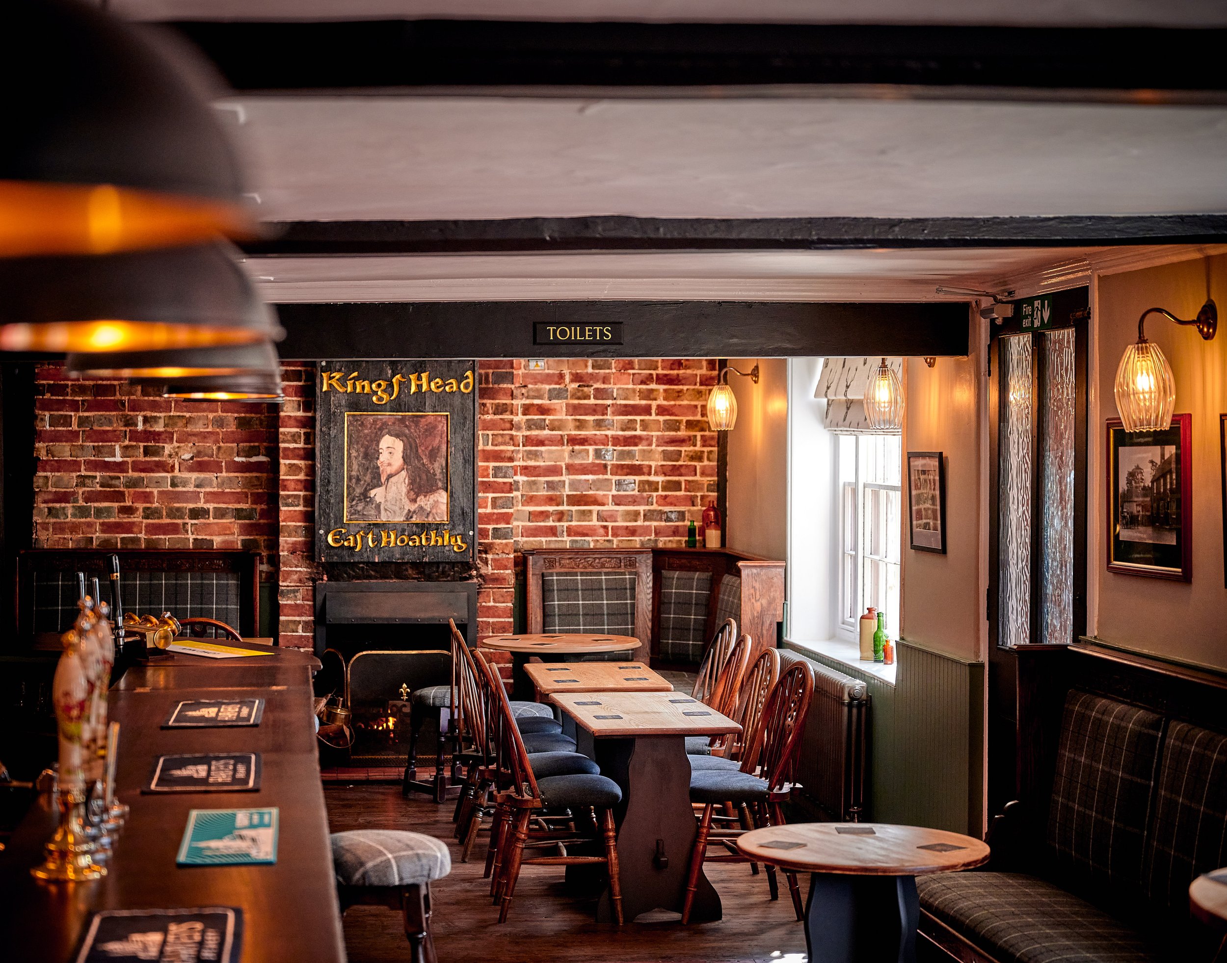 Interior of a pub or restaurant with wooden chairs and tables, brick wall, framed artwork, and hanging lamps, with a sign indicating the toilets and a portrait of a king or historical figure.