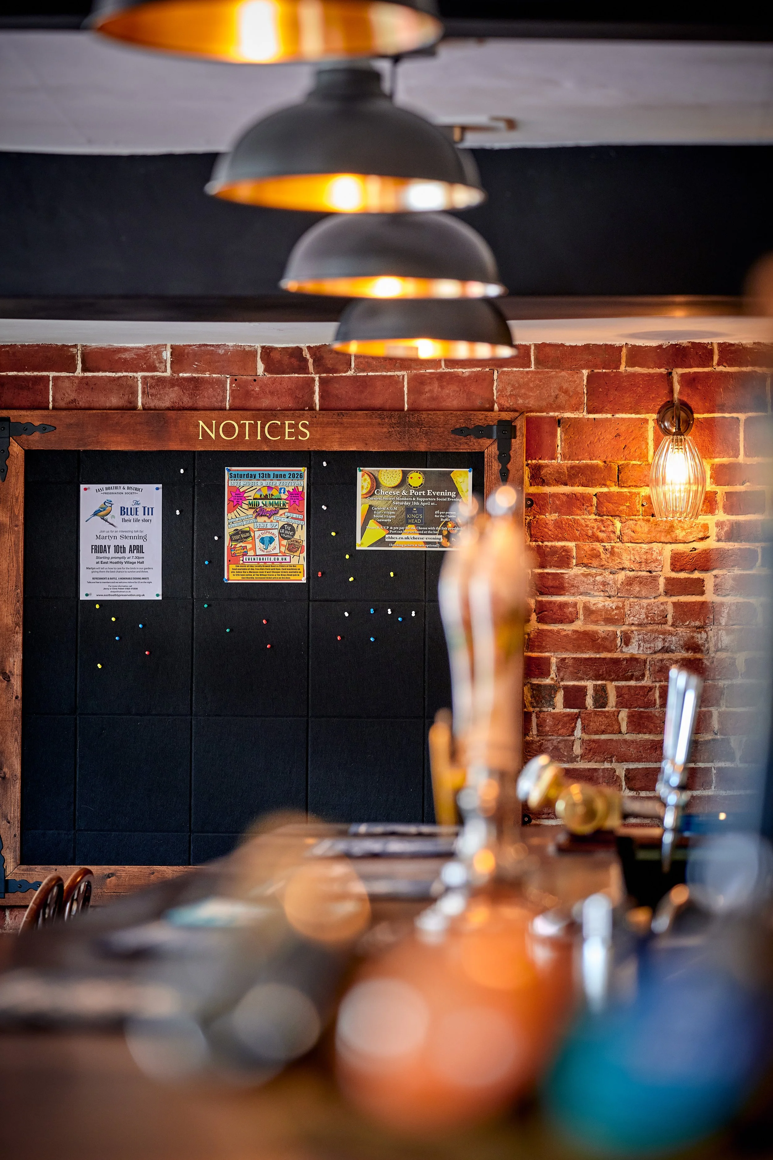 Interior of a pub or bar with black pendant lights hanging from the ceiling, a brick wall with a notice board displaying event posters, and a vintage-style wall sconce with an Edison bulb. The bar counter in the foreground has taps and bar equipment.