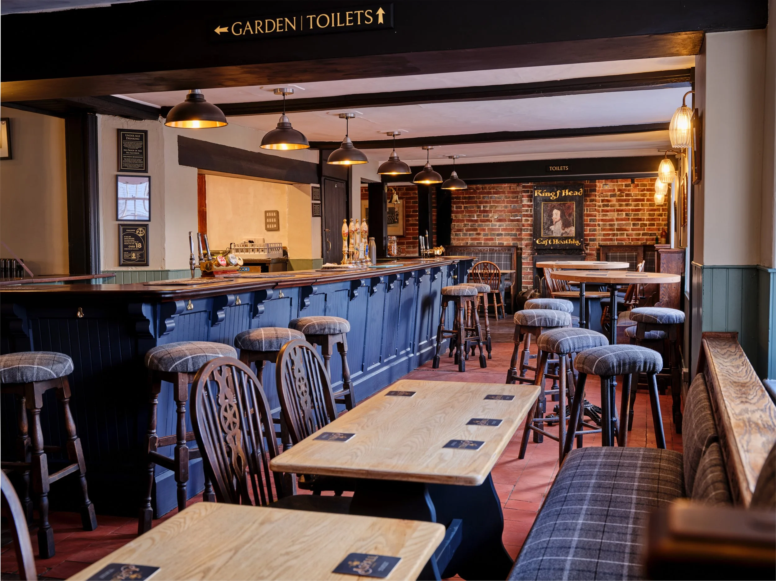 Empty pub or restaurant interior with wooden tables and chairs, a blue bar counter with beer taps, and a brick wall with a framed poster of King Head pub.