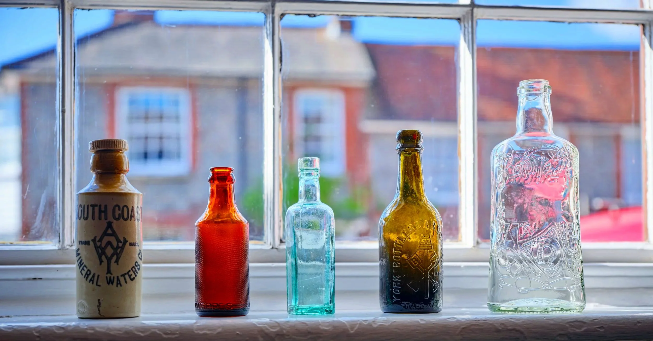 Four colorful glass bottles on a windowsill, with a blurred view of houses outside.