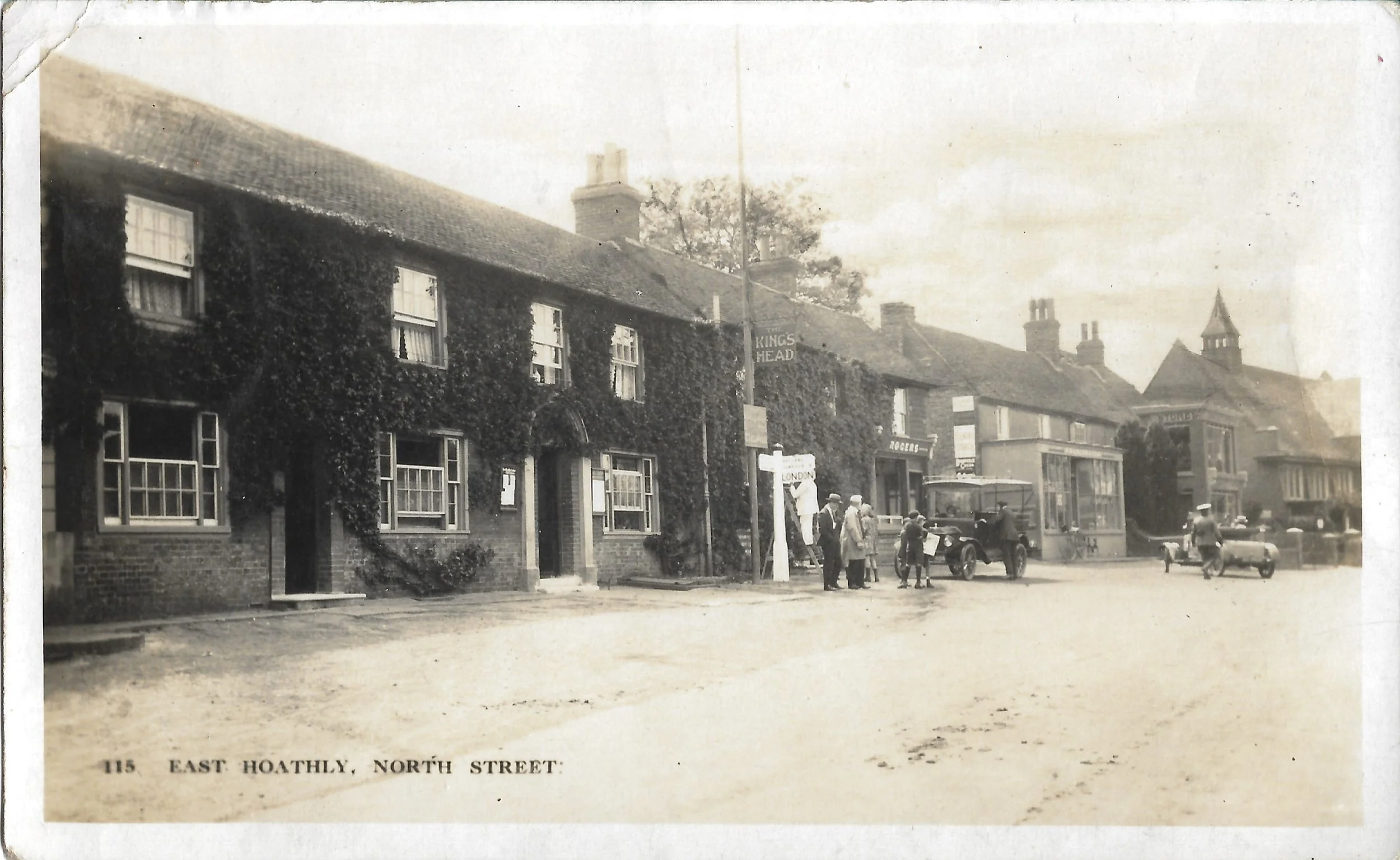 Historic black and white photograph of East Hoathly North Street with building covered in ivy, people, and vintage cars.