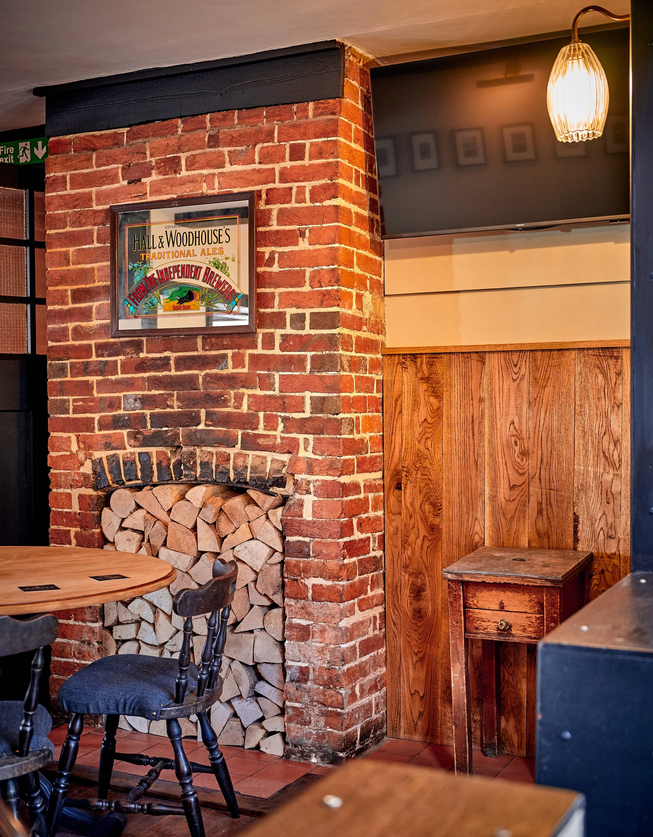 Interior of a restaurant with a brick fireplace with firewood stacked inside. A framed hall and woodhouse sign is hanging on the brick wall. There's a small wooden side table with a drawer to the right of the fireplace. A wooden chair with a black up