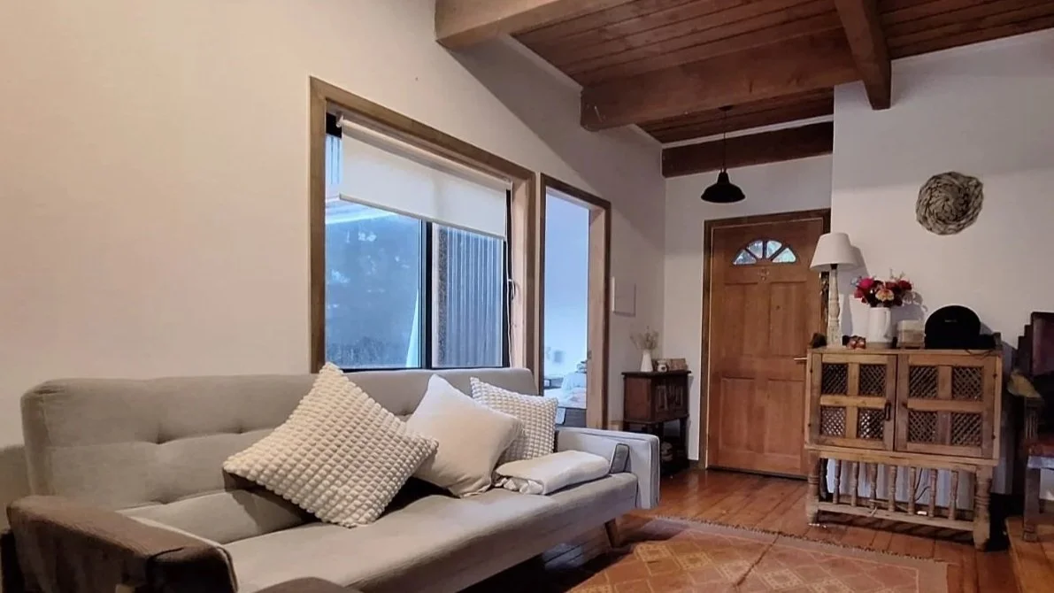 Sala de estar con sillón, ventanas grandes, puerta de madera, lámpara de mesa, mesa pequeña con decoraciones y una pared decorada con objetos, todo en estilo rústico.