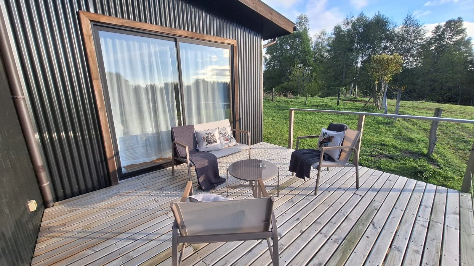 Vista de un balcón de madera con sillones y cojines decorativos, cerca de un campo verde con árboles y una cerca de madera.