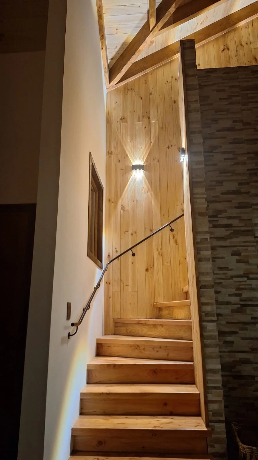 Escalera de madera en un interior con paredes de madera y piedra, con una luz empotrada en la pared iluminando la zona y una baranda en el lado izquierdo.