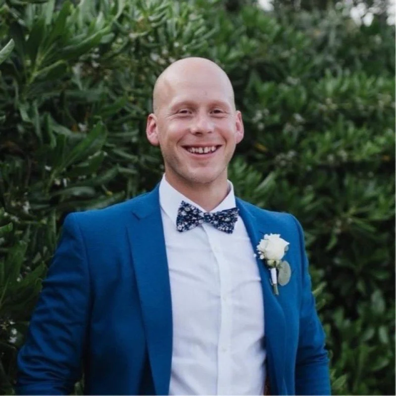 A man in a blue suit with a white shirt and a patterned bow tie, smiling outdoors with green foliage in the background.