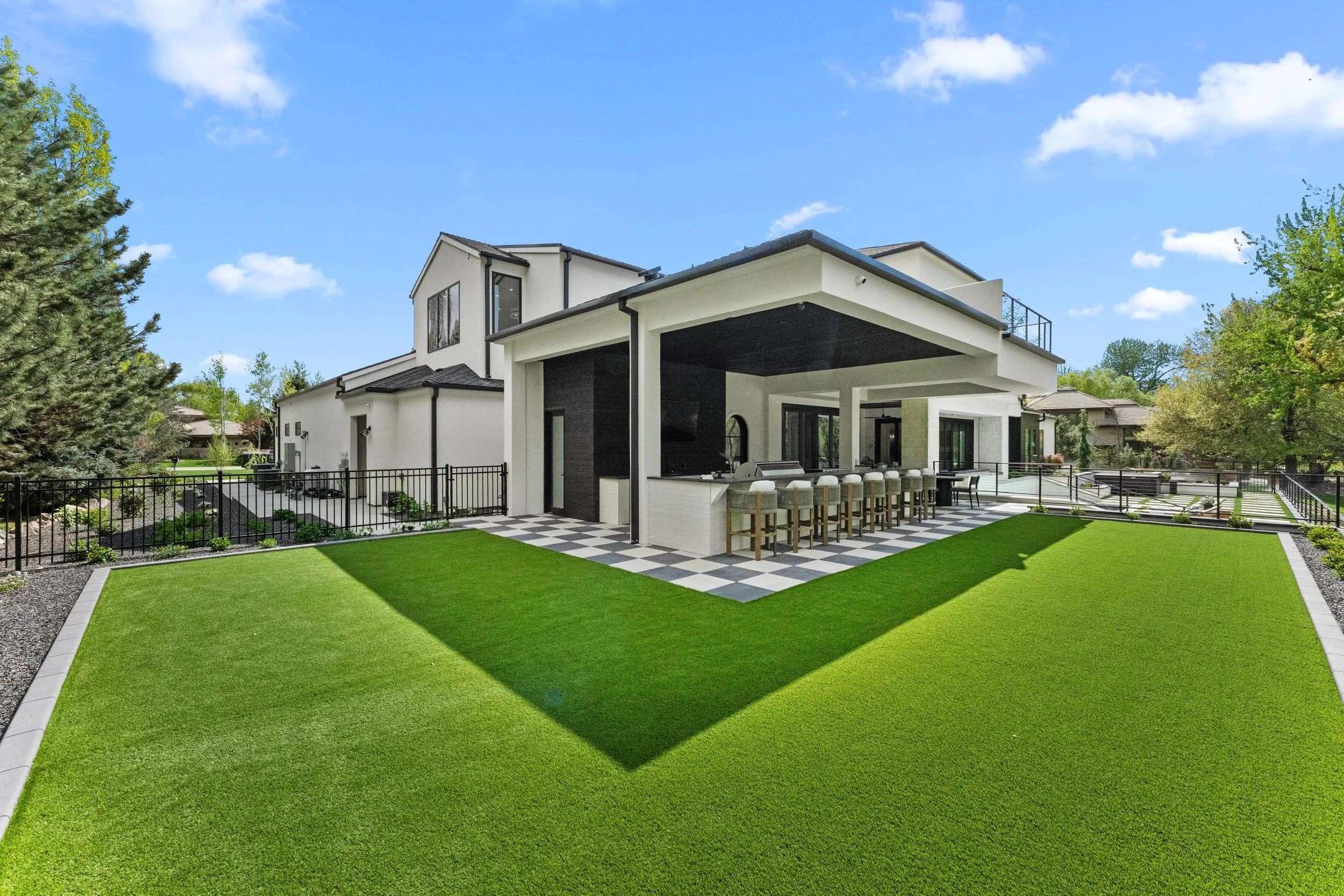 Modern white house with black accents and a spacious backyard featuring a well-maintained green lawn, a checkered patio with barstools, and surrounded by trees and a black fence under a partly cloudy blue sky.