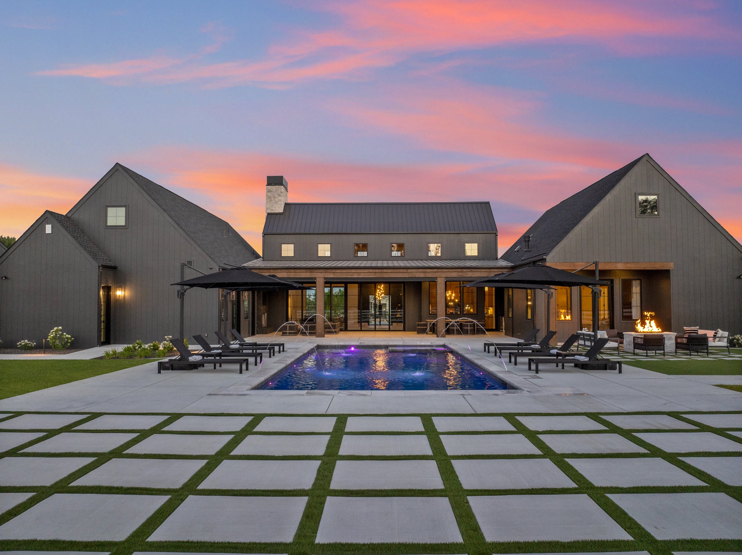 Modern house with dark gray siding, large windows, and outdoor patio area with lounge chairs and firepit, set against a vibrant sunset sky with pink and purple clouds.